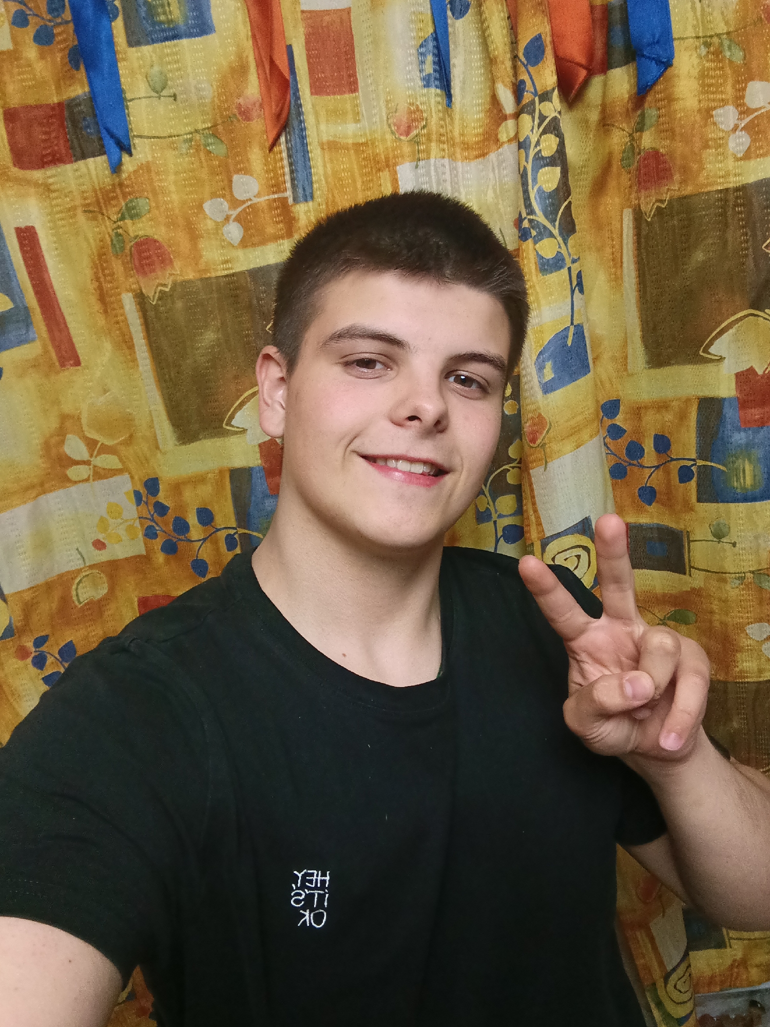 A young man is featured in a close-up selfie, likely taken indoors in Красный Маяк, Russia. He has short, dark brown hair and is smiling broadly, showing his teeth. He is wearing a black t-shirt with white embroidered text on the left chest. Due to the selfie perspective, the text appears reversed, displaying "YƎH ,Ƨ'TI KO", which correctly reads "HEY, IT'S OK". The man is making a peace sign gesture with his left hand, suggesting a casual and friendly demeanor.
The background consists of a colorful, patterned curtain or tapestry. It features a design with abstract shapes and stylized botanical elements in warm tones of yellow, orange, and brown, accented with blue and red details. Several pieces of fabric, appearing as ribbons or strips in blue and orange, hang vertically from the top edge of the curtain. The indoor setting, along with the man's relaxed smile, indicates a casual moment.