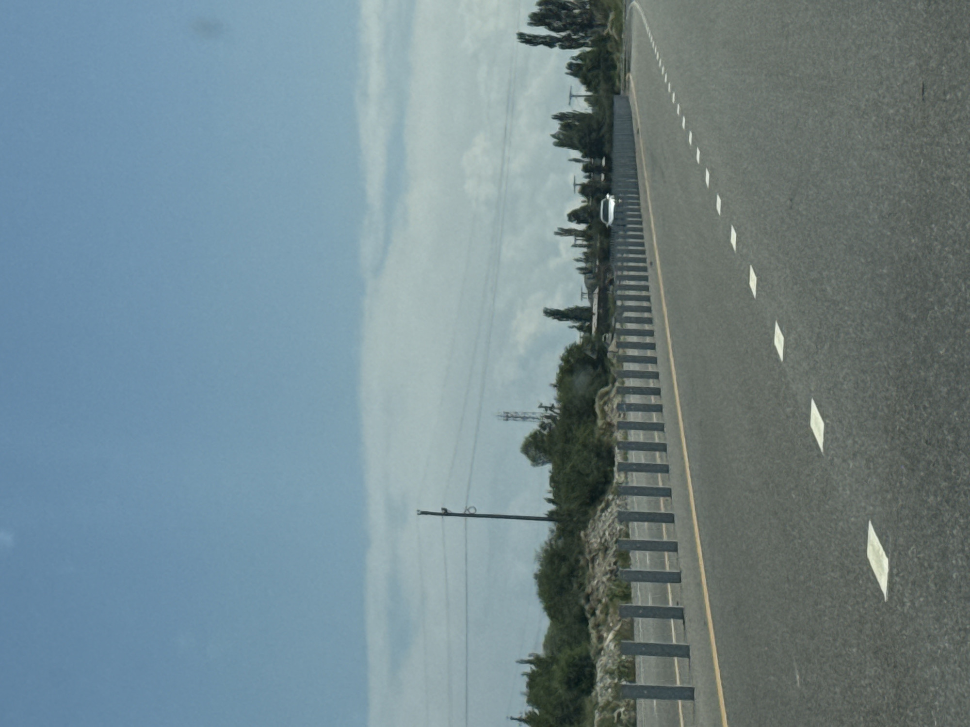 The image depicts a paved highway with intermittent white lane markings, characteristic of a rural or semi-rural setting. The road is bordered on the left by a line of vertical metallic bollards, a safety barrier, and beyond that, a vegetated embankment with scattered trees and shrubbery. A single white vehicle is visible on the road in the distance, beyond the bollard barrier. In the background, a power line pole with visible wires stretches across the frame, alongside more trees and what appears to be a radio tower. The sky is predominantly blue with scattered clouds, indicating daytime. This scene is consistent with the landscape found in and around Kara-Talaa, Kyrgyzstan, known for its open terrain and infrastructure supporting transportation routes. There are no people visible in the image.