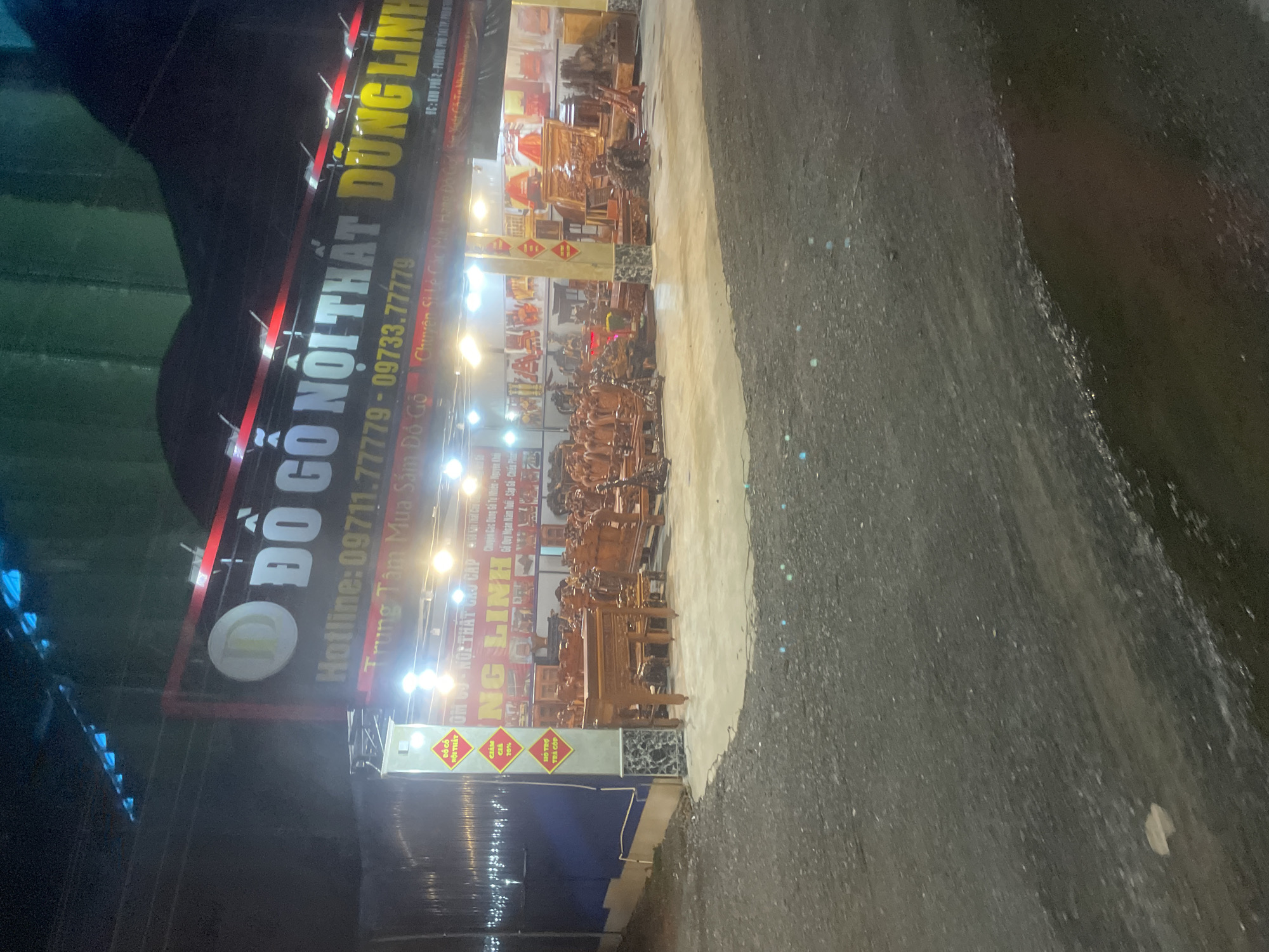The image shows a nighttime view of a furniture store in Binh Thuan Ward, Vietnam. The store, named "ĐỒ GỖ NỘI THẤT DŨNG LINH," is illuminated by bright lights. A large sign above the store displays the name and contact information: "Hotline: 09711.77779 - 09733.77779." The store front is filled with various wooden furniture pieces, including chairs, tables, and intricate carved pieces. Some of the furniture is displayed on a paved outdoor area in front of the store, while more is visible inside through the open facade. Yellow banners with red text are attached to a pillar on the left side of the entrance, indicating "ĐỒ GỖ NỘI THẤT" and "GIẢM GIÁ 10%". The ground in front of the store is a rough, dark surface, possibly gravel or asphalt, with scattered small reflective elements, possibly due to moisture or lighting. The surrounding environment is dark, suggesting it is late at night.