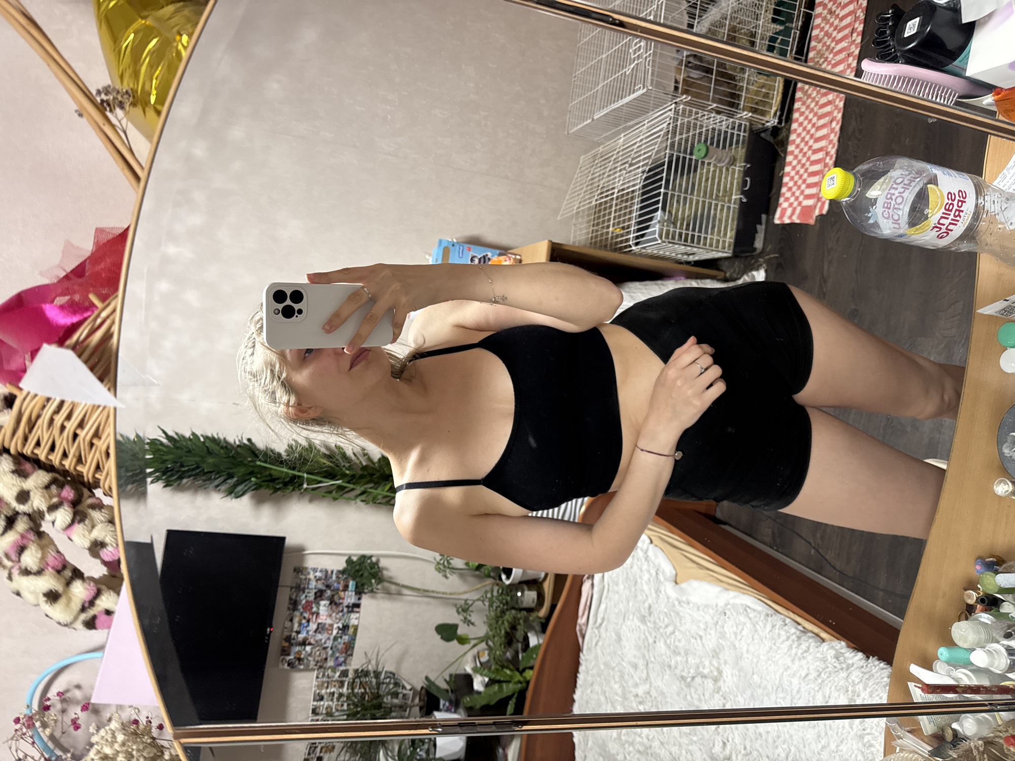 A young woman with blonde hair is taking a mirror selfie in a room. She is wearing a black sports bra and black shorts. Her reflection shows her holding a white smartphone in her right hand. The background of the room includes a bed with a fluffy white blanket, a wooden dresser, several potted plants, and animal cages. There are also various items on the dresser in the foreground, including a bottle of water and toiletries.