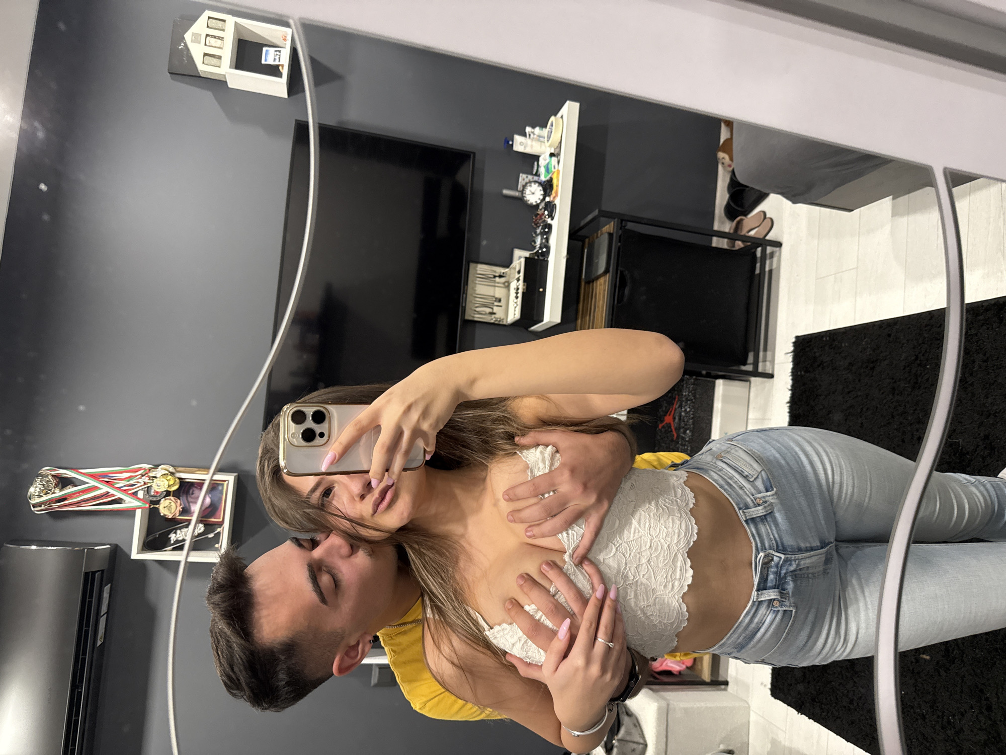 A couple is taking a selfie in a mirror. The woman is wearing a white lace top and light blue jeans. Her hands are on her chest, and her boyfriend's hands are on top of hers. The background shows a modern apartment with gray walls, a large flat-screen TV, and various decorative items on shelves. There are also Olympic medals and a picture. The location appears to be a modern apartment. Given the style and appearance of the apartment, it is likely located in an urban area, specifically in Sofia, Bulgaria.