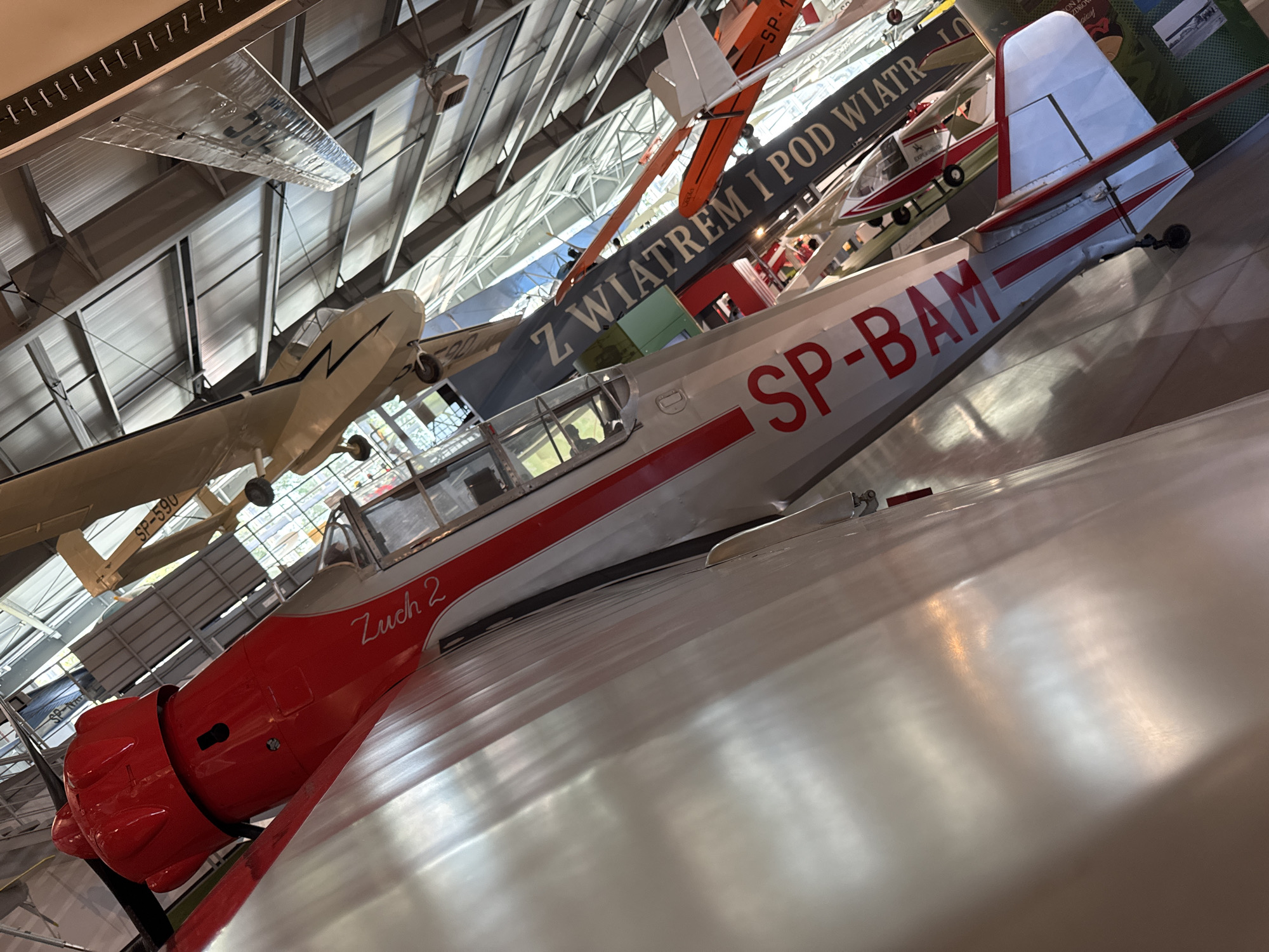 This is a close-up, tilted, eye-level shot of a white and red airplane on display indoors, likely in a museum. The plane is the primary subject and is positioned diagonally in the frame, with its nose on the left and tail on the right. The fuselage has a prominent red stripe running along its length, and the word "Zuck 2" is written in a cursive font on the side of the cockpit area. The registration number "SP-BAM" is clearly visible in large red letters on the fuselage. The cockpit canopy is visible, and the plane has landing gear.
In the background, other aircraft are suspended from the ceiling, suggesting an aviation museum setting. The ceiling itself is made of corrugated metal and exposed beams, with large skylights or windows allowing natural light to filter in. Reflections are visible on the metallic surface of the airplane's fuselage, indicating a polished finish. The overall environment appears to be a spacious indoor exhibition hall.