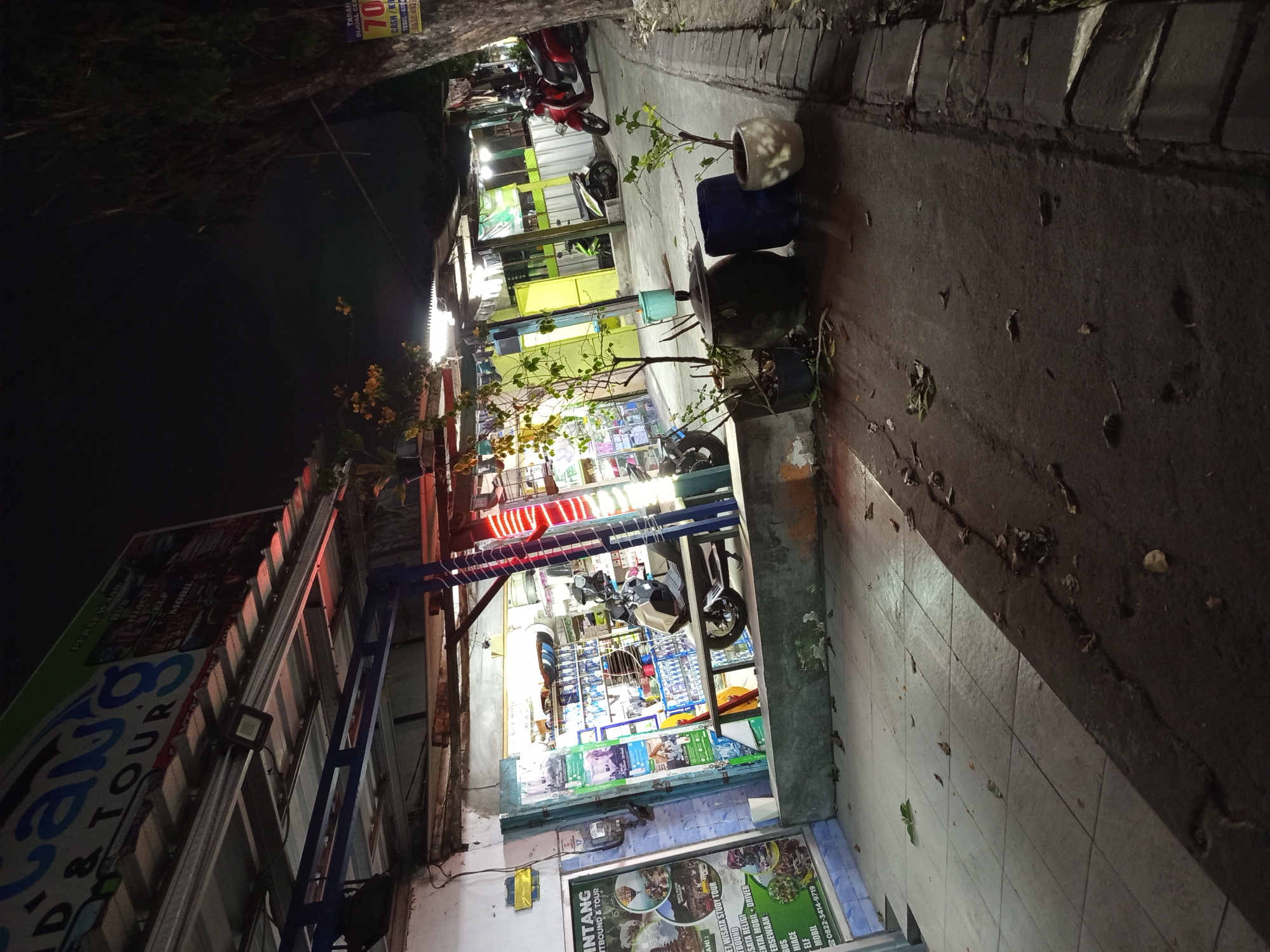 The image depicts a street scene at night in Sruni, Indonesia, with a focus on a small storefront and the adjacent sidewalk. The storefront appears to be a shop selling various goods, with shelves stocked with items visible through a glass door. A white and beige scooter is parked in front of the shop. Red neon lights are displayed on the side of the building. Outside the shop, there are several potted plants, two large dark-colored containers, and a lighter-colored pot. A red motorcycle is parked further down the street. Fallen leaves are scattered on the sidewalk and street. A tree trunk is visible on the right side of the image, with a poster attached to it advertising "TANAH DIJUAL" (Land for Sale) with a price of "70" and a phone number. The overall atmosphere is that of a dimly lit, bustling commercial area at night.