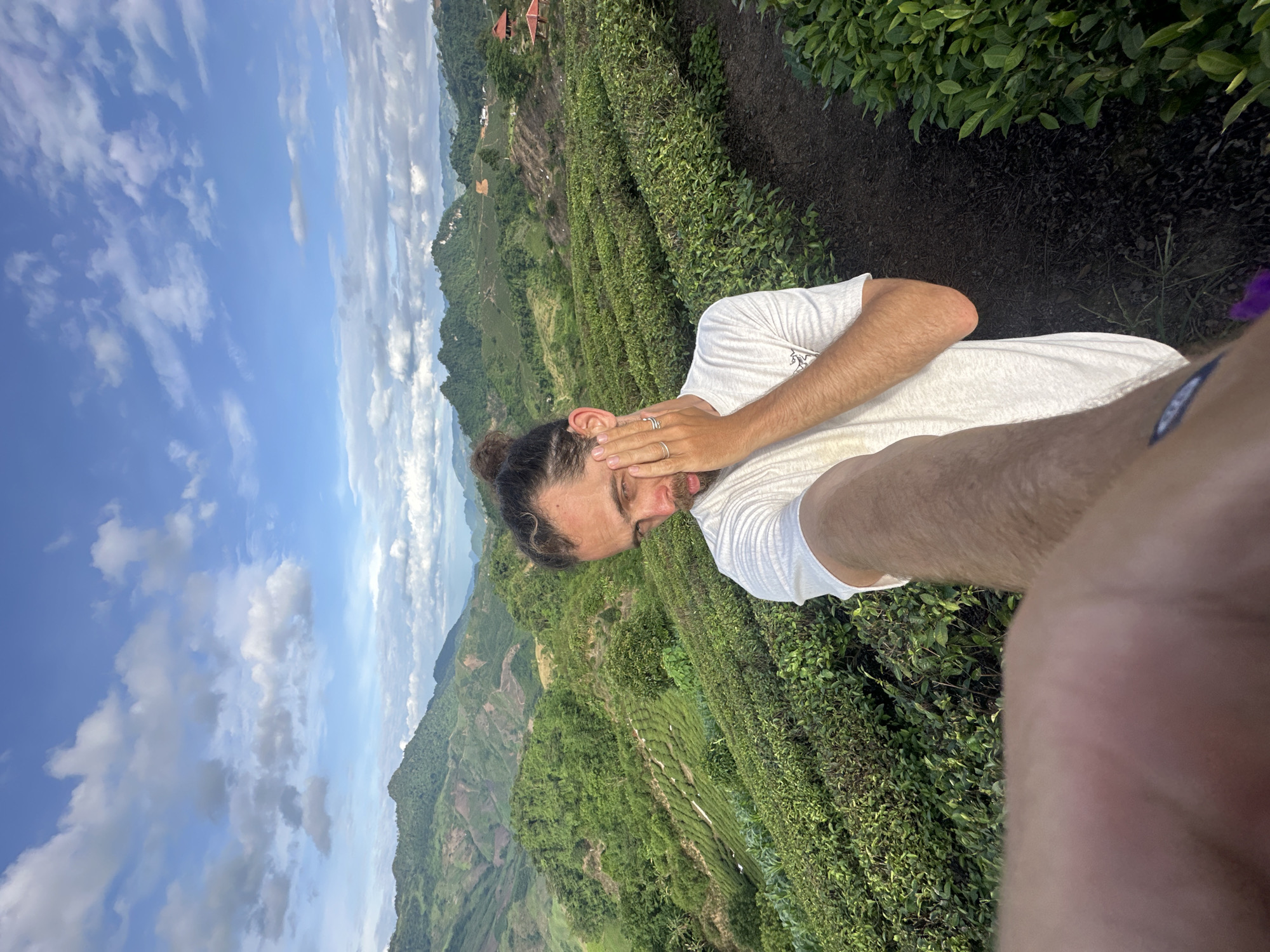 A man is taking a selfie in a tea plantation in the mountains of To Mua, Vietnam. The man has his hair pulled back in a bun and is wearing a white t-shirt. He is covering his eye with his hand. The tea plantation is on a hillside, and in the background, there are more hills and mountains. The sky is blue with some clouds.