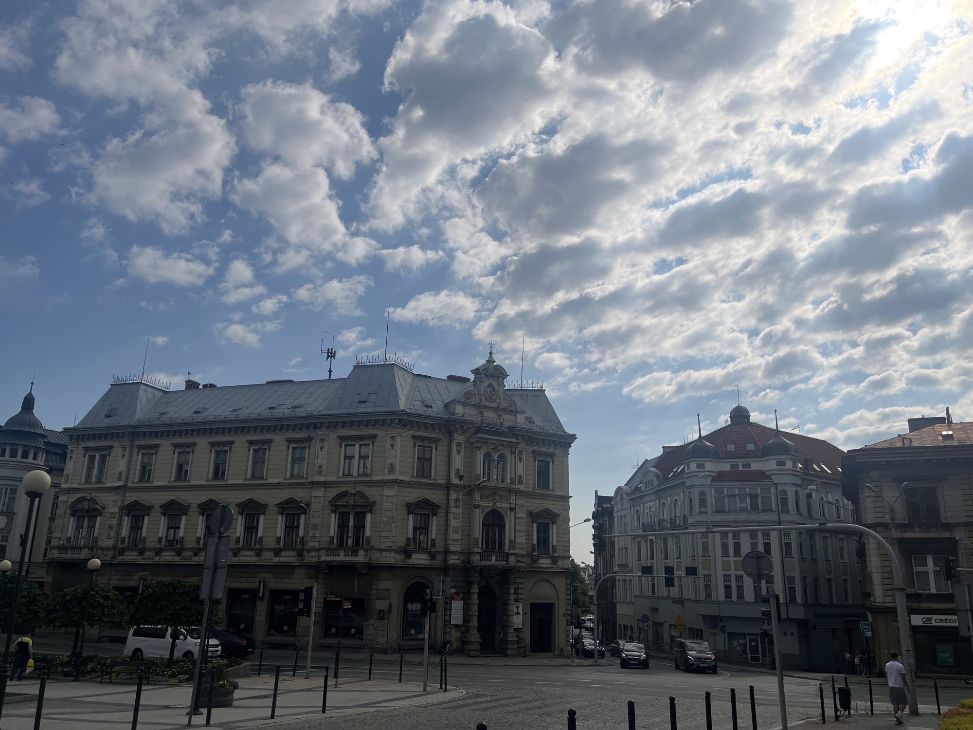 In an urban environment, a street scene unfolds under a partly cloudy sky with a bright sun peeking through. Two prominent buildings dominate the right side of the image. The building on the left is a tan, multi-story structure with a grey, sloped roof adorned with dormer windows and decorative elements along the facade. It has a symmetrical design with numerous windows and balconies. To its right, a lighter blue and grey building features a distinctive curved facade and a red, domed roof with smaller domes. This building also has multiple stories with numerous windows. Cars are visible on the street, suggesting it is a thoroughfare. One car, a white van, is parked on the left side of the image. A few pedestrians are present, including a person in a yellow shirt walking with a yellow bag on the left, and individuals on the sidewalk near the right building. The street is paved and bordered by bollards and streetlights. The overall impression is that of a city square or intersection in a European city.