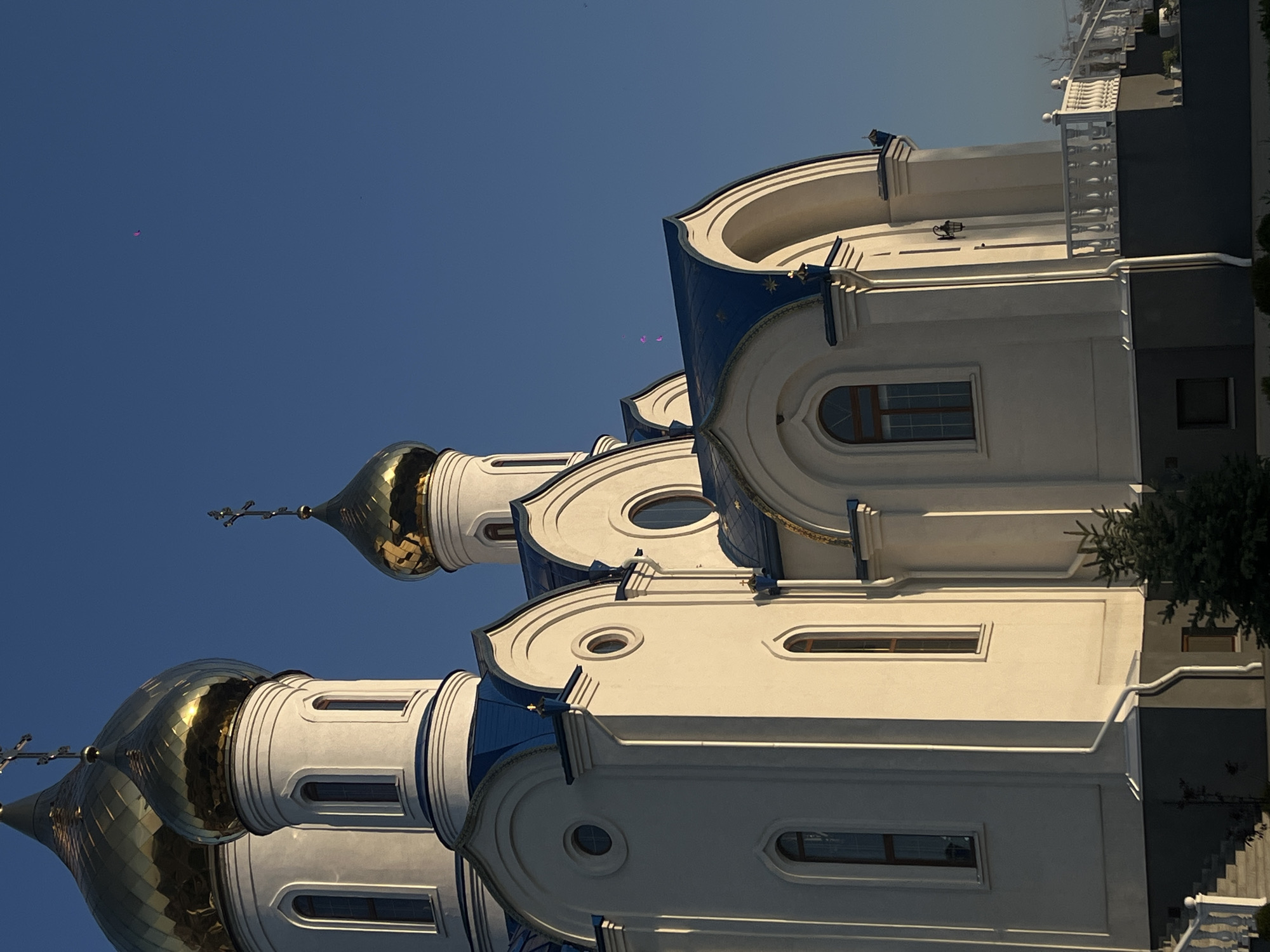 A majestic Russian Orthodox church, predominantly white with striking blue accents and golden domes, stands prominently against a clear, vibrant blue sky. The image captures a portion of the church building, angled upwards, with its ornate architecture illuminated by bright sunlight.
The church features multiple levels and sections, characterized by smooth, white walls and intricate decorative moldings. Several windows are visible; some are tall and arched, others are small and circular, adding to the building's detailed design. The roofs and specific architectural elements are painted a rich blue, with some sections appearing to have subtle star patterns.
Two large, gilded onion domes, characteristic of Orthodox Christian architecture, gleam brightly in the sunlight, reflecting a warm golden hue. Each dome is topped with a slender spire and an ornate cross. A third, smaller golden dome is partially visible in the background.
To the right of the church building, a white balustrade or railing is present, suggesting a balcony or an elevated area. A dark-colored section of an adjacent building and a small evergreen tree or bush are also visible on the far right.
The sky is a uniform, deep blue, indicating a bright and cloudless day. A few small, faint pink or purple specks are visible in the upper left quadrant of the sky, likely lens flares, along with some tiny dark specks that could be distant objects or dust. The strong sunlight casts clear shadows, highlighting the building's architectural depth and texture. No human activity or visible text is present in the scene. The setting appears serene and grand.