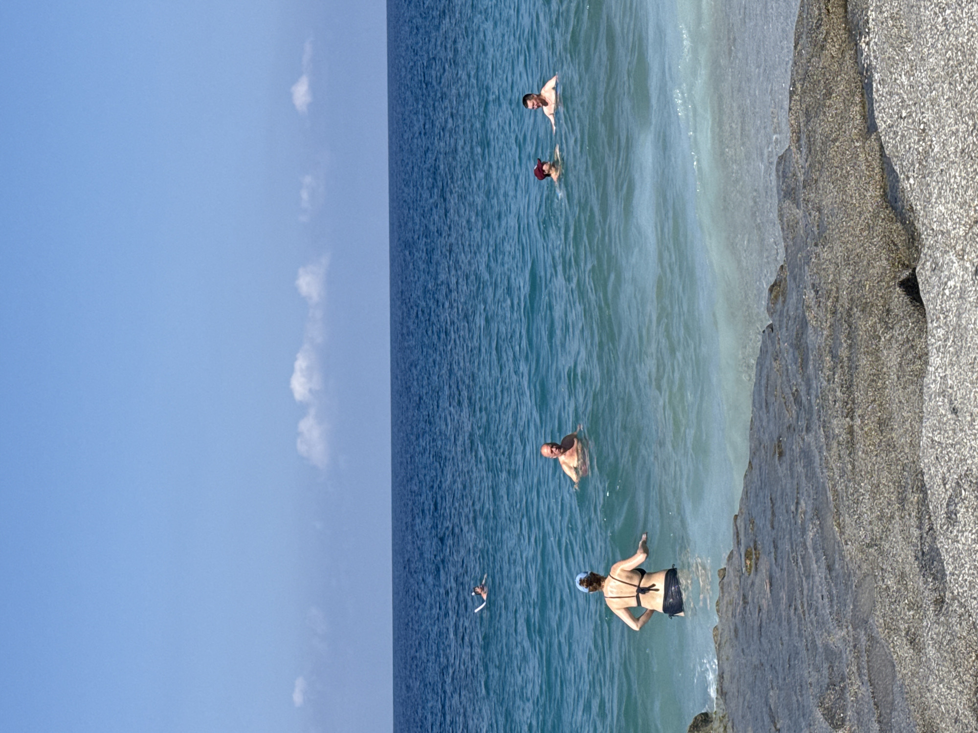 The image depicts a sunny day at a beach in Alanya, Turkey. Several people are seen bathing in the clear blue water. In the foreground, a woman wearing a black bikini and a blue cap is standing in the shallow water, facing away from the camera. Further out in the sea, a man is swimming, and two other individuals are also visible in the water. The background features the vast expanse of the blue sea meeting the horizon under a clear blue sky with a few scattered white clouds. A rocky shoreline is present in the immediate foreground.