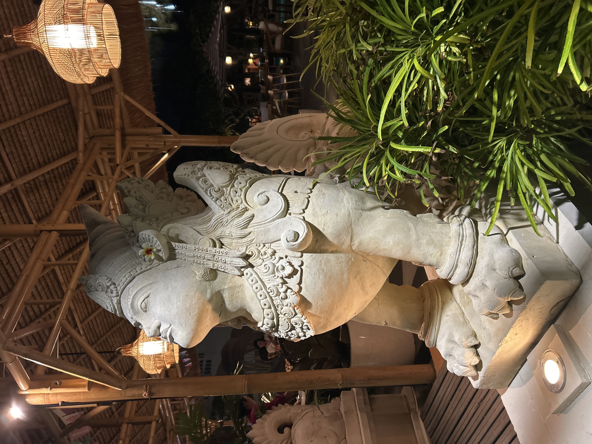 The image depicts a stone sculpture of a winged creature, possibly a mythical being from Balinese culture, positioned outdoors in what appears to be a restaurant or dining area with a thatched roof and bamboo structures. The sculpture is illuminated by warm, ambient lighting, including woven pendant lamps and ground-level spotlights. Lush green foliage is visible in the foreground and to the right of the sculpture. In the background, there are glimpses of tables and chairs, suggesting a seating area, and silhouetted figures of people. The overall ambiance is tropical and perhaps a bit exotic. The scene is likely located in Denpasar, Indonesia, given the cultural context of the sculpture and the architectural style.