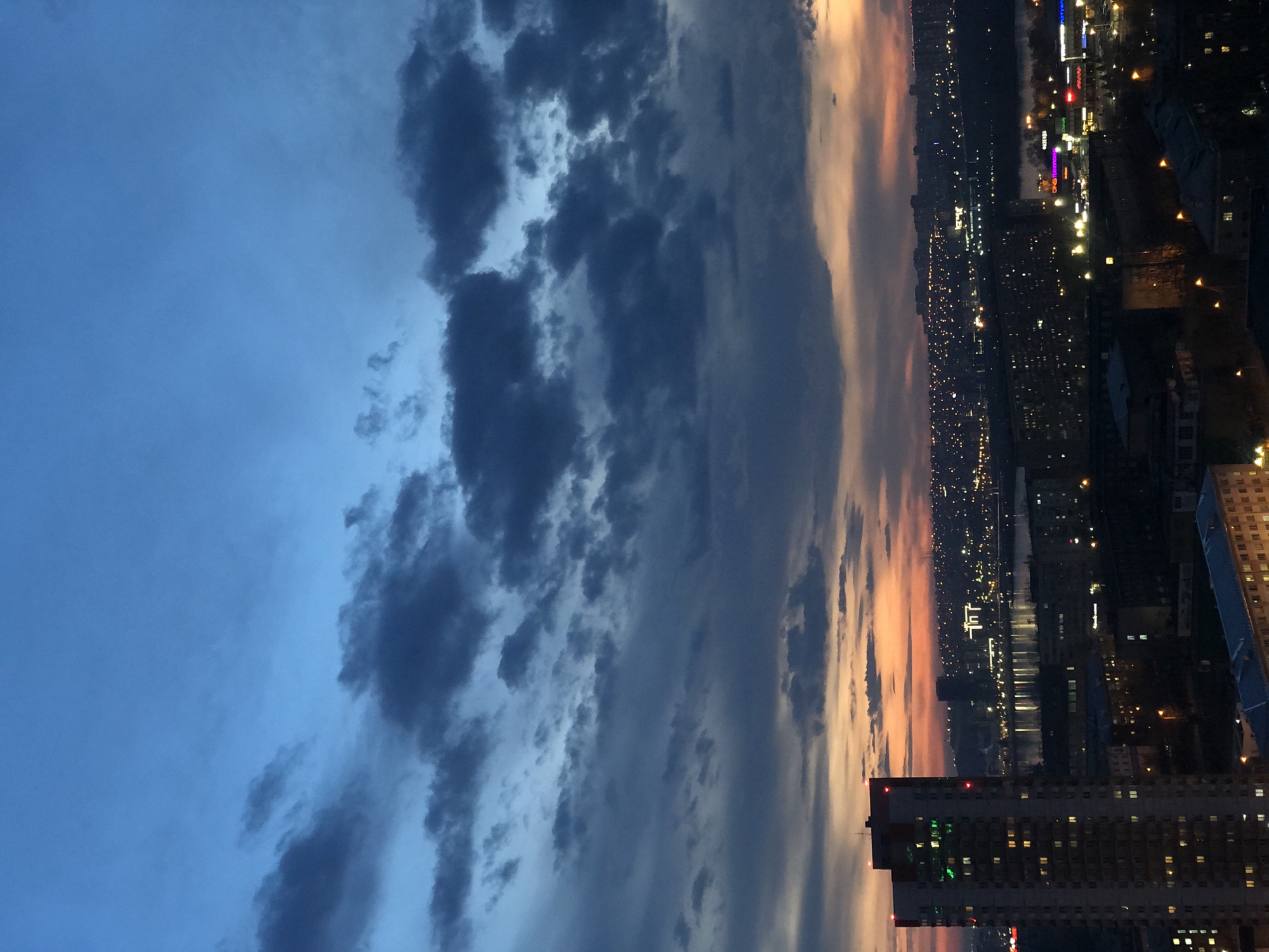The image presents a panoramic high-angle view of the city of Krasnoyarsk, Russia, captured during twilight or early night. The expansive sky, occupying roughly half of the frame, transitions dramatically from a deep blue on the left to warm orange and pink hues on the right horizon, suggesting a recent sunset. Dark, textured clouds are prominent in the central sky, adding depth to the scene.
Below the dynamic sky, the urban landscape of Krasnoyarsk stretches out, illuminated by countless city lights. A wide, dark body of water, likely the Yenisei River, horizontally cuts through the city, reflecting some of the distant lights. Numerous buildings, including residential and commercial structures, line the riverbanks and populate the foreground. Many windows are lit, creating a shimmering pattern across the cityscape. In the right foreground, a tall building with illuminated windows and red lights on its roof stands out.
Visible text on commercial signs includes "DNS" and "KRASSTROY" on a brightly lit building in the mid-ground, and "AZIMUT" on a more distant structure. The scene is static, capturing the city in a tranquil yet vibrant state as night falls.