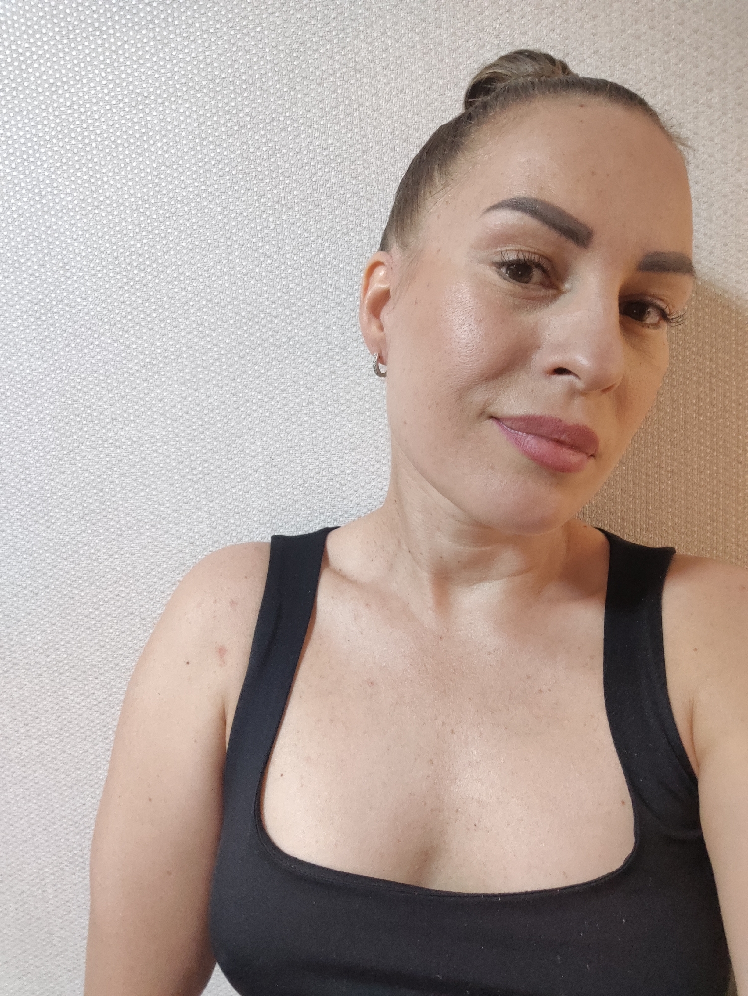 This is a medium close-up, selfie-style photograph of a woman in Almaty, Kazakhstan. The woman has her hair pulled up in a bun and is wearing a black tank top. She has a light dusting of freckles and appears to be wearing light makeup, including lip gloss. She is looking towards the camera with a slight smile. The background is a textured, light-colored wall, suggesting an indoor setting. The lighting appears to be even and soft, possibly from natural light or diffused artificial light, and there are no obvious shadows indicating a specific time of day. The overall mood of the image is casual and relaxed.