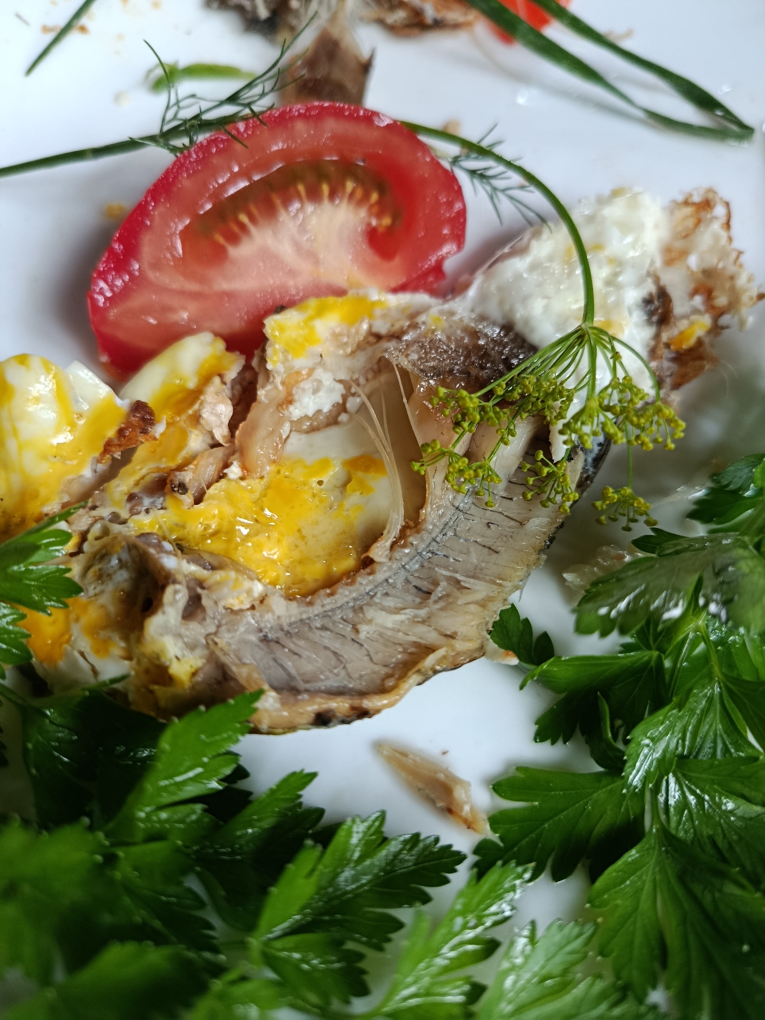 A close-up, overhead shot showcases a white plate with a serving of food. The main subject is a piece of cooked fish, its skin slightly browned, nestled within what appears to be baked eggs, with visible portions of cooked egg white and a vibrant yellow yolk. A slice of red tomato sits to the upper left of the fish, and a sprig of fresh dill with tiny yellow flowers garnishes the dish. Lush green parsley leaves frame the bottom and right sides of the plate. The background is a plain white surface, presumably the plate itself. The image is well-lit, highlighting the textures and colors of the food. There are no people or any indication of the location being in the city of Верея, Russia.