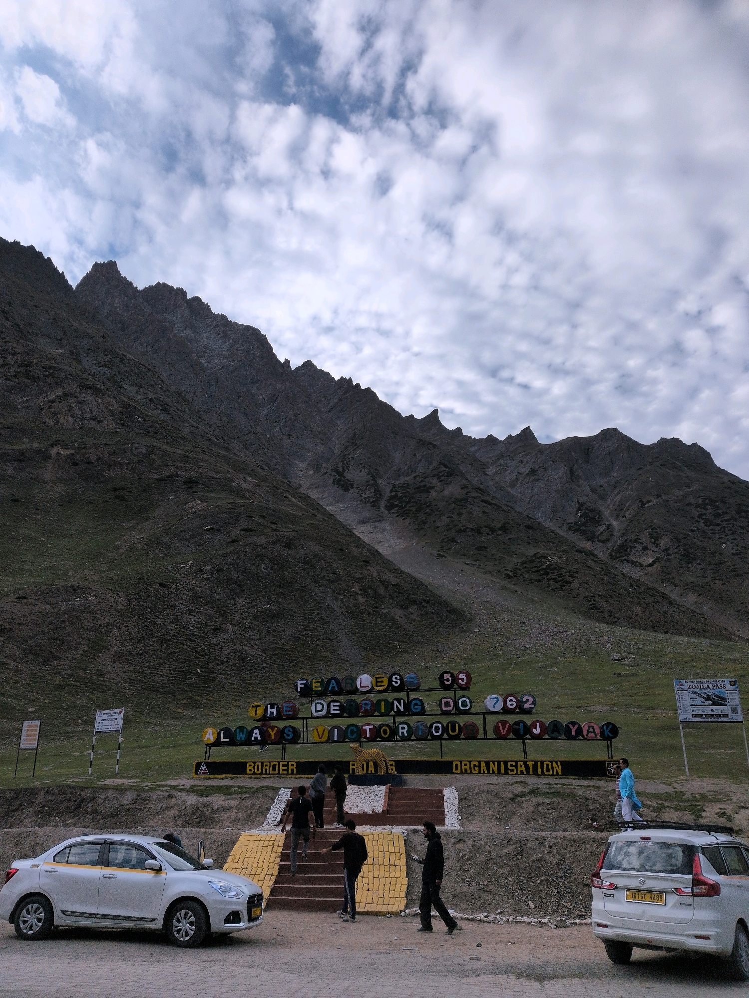 The image captures a scenic view of a mountainous region in India, with a prominent sign that reads "FEARLESS 55 THE DEEPING DO 762 ALWAYS VICTORIOUS VIJAYAK BORDER ORGANISATION ZOJILA PASS". The sign is composed of colorful, individual letters and numbers mounted on a raised platform. Several people are visible near the sign, some walking up a yellow and brown staircase, and others near vehicles.
In the foreground, a white sedan is parked on the left, and a white SUV is parked on the right. The SUV has a license plate that reads "JK16C 4489". The environment is characterized by rugged, green and brown mountains under a cloudy sky. A smaller sign is visible on the right side, which appears to provide information about Zojila Pass. The overall scene suggests a location along a border road or mountain pass, likely a tourist or transit point. The weather appears to be overcast, with visible clouds in the sky.