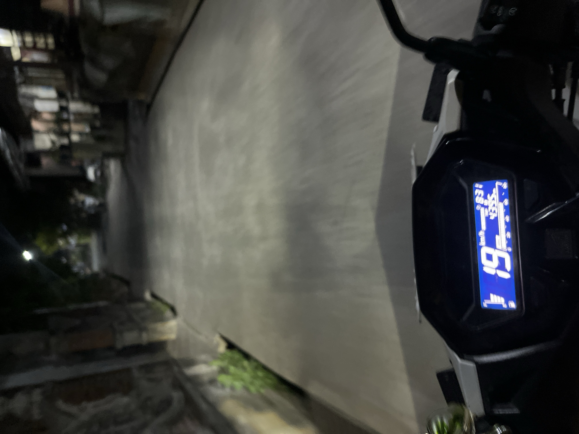 The image is a first-person perspective shot taken at night, likely from the viewpoint of someone riding a scooter or motorcycle. The focus is on the digital display of the vehicle's dashboard, which shows a speed of "19 km/h" and other indicators. Beyond the dashboard, a dimly lit street is visible, lined with buildings and vegetation. A street lamp illuminates a section of the road. The general atmosphere suggests a quiet, residential area in Karangrandu, Indonesia, during nighttime. No people are clearly identifiable in the image.