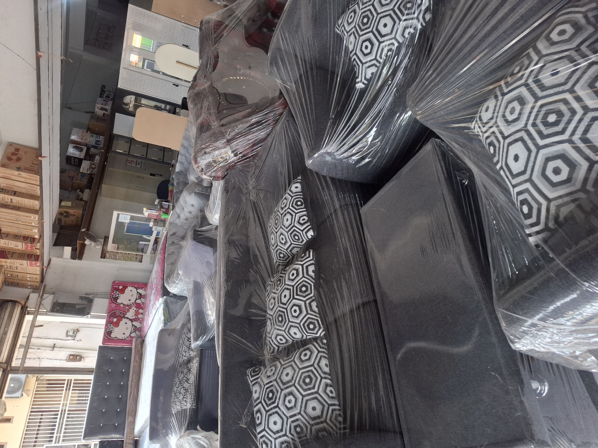 A furniture store in Banjarmasin, Indonesia, is filled with various upholstered items, primarily sofas and beds, all covered in transparent plastic wrap for protection. The lighting is somewhat dim, suggesting an indoor environment, possibly a warehouse or showroom. In the background, shelves stocked with boxes are visible, along with what appear to be metal cabinets and a closed roll-up door. The furniture includes a bed with a headboard featuring Hello Kitty characters, multiple sofa sets in dark upholstery with geometric patterned cushions, and an armchair. The overall impression is one of a retail space preparing for or undergoing inventory or display.