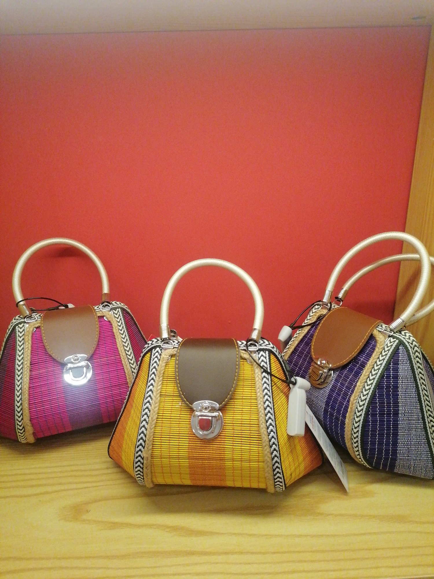 Three vibrantly colored, handmade handbags are prominently displayed on a light-toned wooden surface against a solid reddish-orange wall. The bags, likely crafted from woven textile, showcase distinct patterns and color schemes. The bag on the left features magenta and pink stripes, the central bag is in yellow and orange, and the bag on the right displays purple and blue hues with gray and white accents. Each handbag has light, curved handles, a brown leather-like flap with a silver-toned turn lock closure, and decorative braided borders. Two of the bags, the central and rightmost ones, have unreadable white tags attached, possibly price or brand labels. A vertical wooden panel or beam is visible on the far right of the image, suggesting an indoor setting such as a shop or market stall. The artisanal style, with its traditional woven patterns and bright colors, is characteristic of local crafts found in Sa Pa Ward, Vietnam, reflecting the textile heritage of the region. The scene is brightly lit, indicating an interior environment.