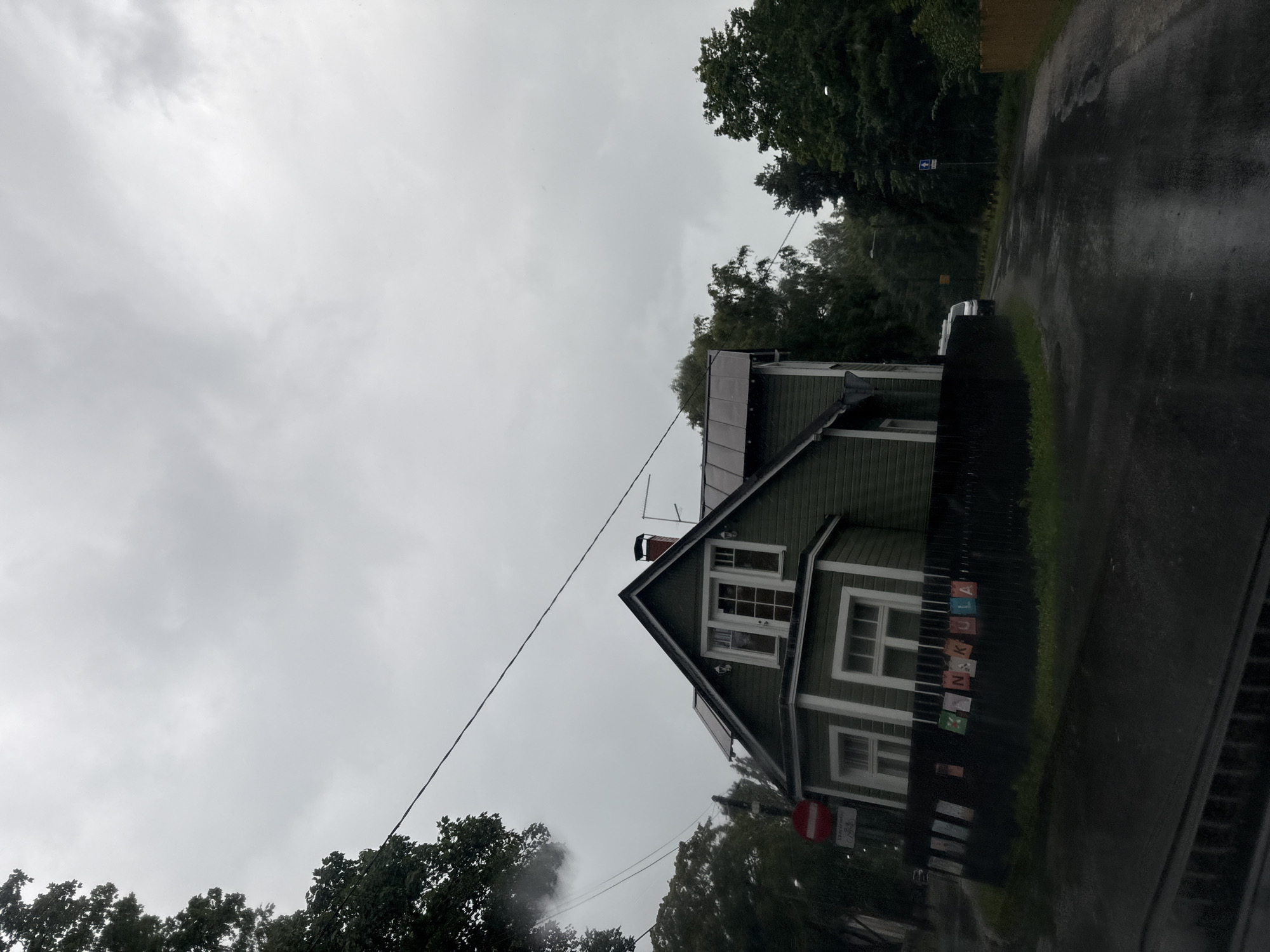 The image depicts a cloudy day in Tallinn, Estonia, with a residential house and a street. The house is a two-story, dark green wooden structure with white trim and windows. It has a steep, dark roof and a chimney. In front of the house, a black wooden fence has colorful letters attached, spelling out "HANKULA". To the left of the house, a red and white circular sign is visible. The street in front of the house is wet and appears to have been recently rained on. Trees line the street, and a white van is parked further down the road. The sky is overcast with thick gray clouds.
