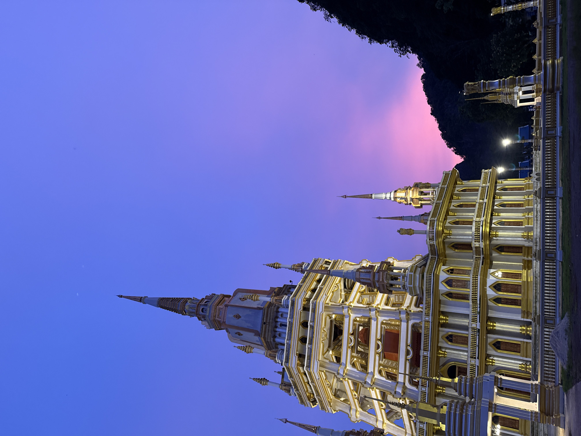 A wide-angle, low-angle outdoor shot captures the Wat Tham Khao Phra temple at dusk in Ban Khao Tang, Thailand. The temple is a multi-tiered, ornate structure with white and gold accents, featuring numerous spires and detailed architectural elements. It is illuminated by artificial lights, highlighting its intricate design. A gradient sky of deep blue at the top transitions to vibrant pink and purple hues near the horizon, casting a dramatic glow. To the right, a dark, tree-covered mountain range rises steeply, silhouetted against the colorful sky. The foreground shows a paved area with a low, decorative fence and a small pile of sand. No people are visible in the frame.