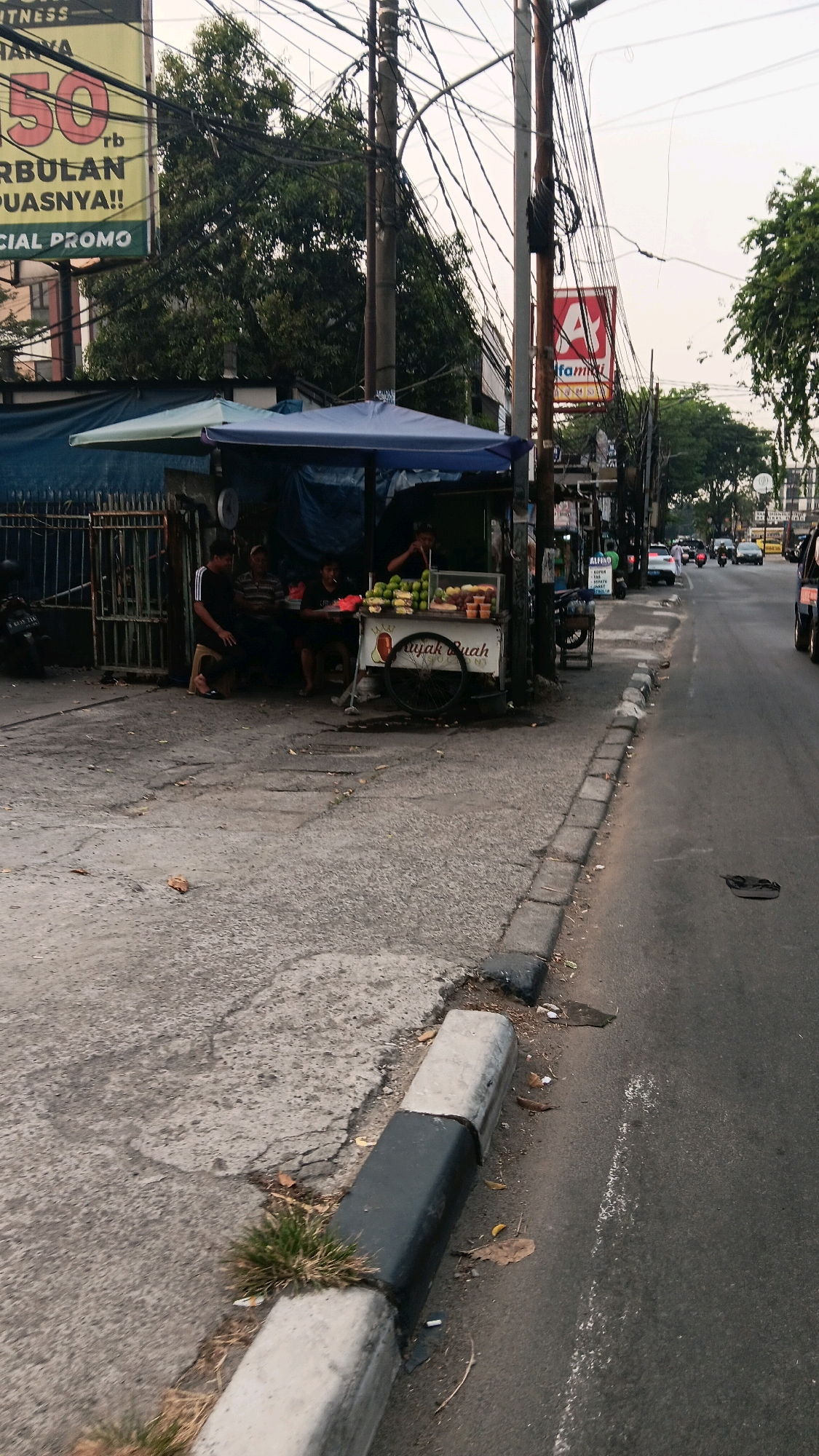 The image shows a street scene in South Jakarta, Indonesia. In the foreground, a sidewalk curb is visible, with some grass growing in the cracks. The street is paved and has a white line painted on it. On the left side of the street, there is a food stall selling fruit juices. Several people are sitting and standing around the stall. Behind the stall, there are buildings and trees. In the background, there is a busy street with cars and motorcycles. There are also many utility poles with tangled wires overhead. A sign for "ALFAMART" is visible on the right side of the image. The overall scene depicts a typical urban environment in South Jakarta, with a mix of commercial activity and everyday life.