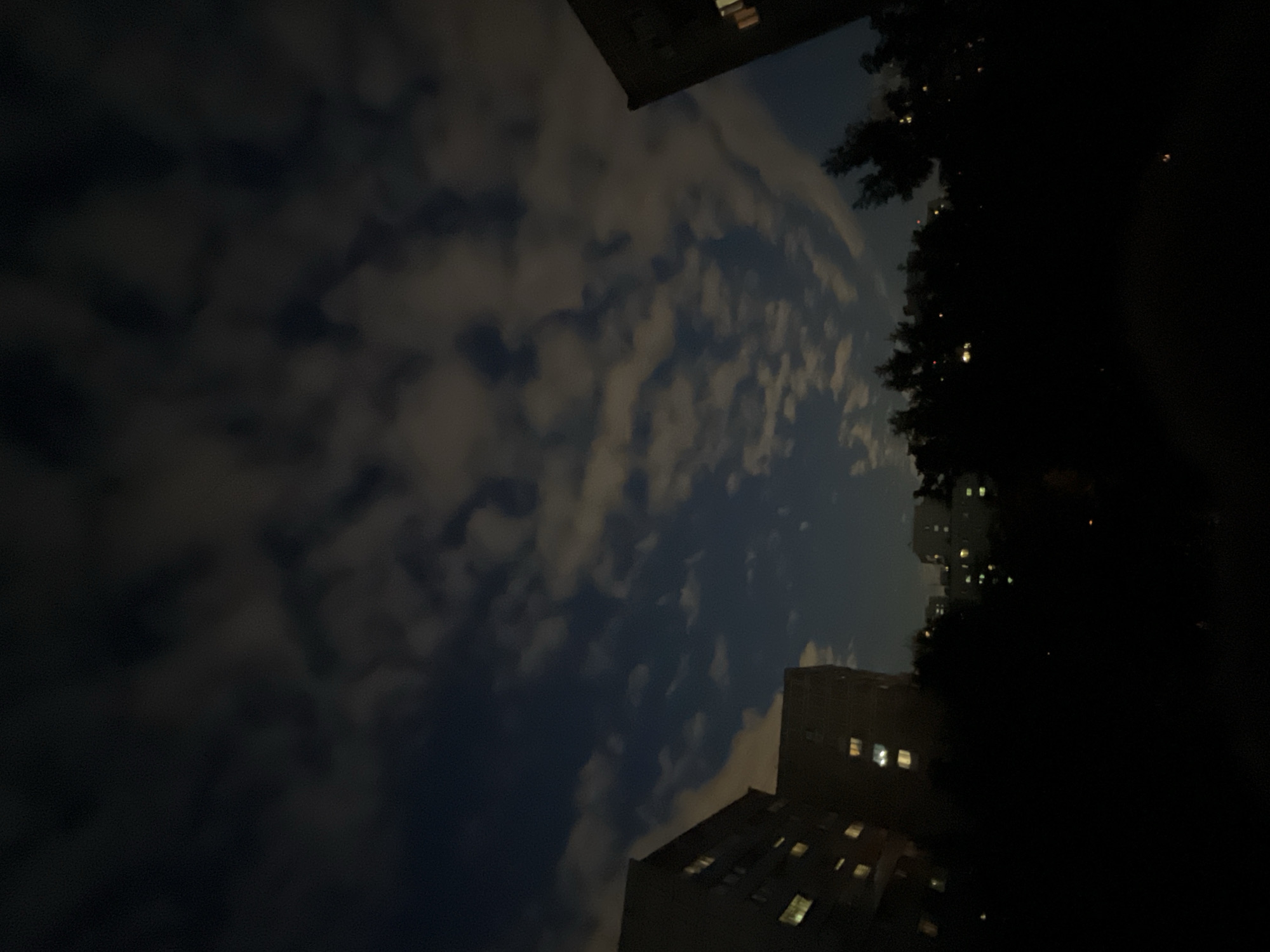 The image captures a night scene in the city of Khimki, Russia, characterized by a dark, cloud-filled sky and the silhouettes of buildings and trees. The sky is a deep blue, partially obscured by scattered clouds that reflect some light, giving them a luminous quality. In the foreground, the dark outlines of trees dominate the lower portion of the frame. Behind the trees, several multi-story apartment buildings are visible. Their windows are illuminated, indicating occupancy. To the left, two distinct buildings stand out, with multiple lit windows on their facades. To the right, the corner of another building is partially visible, also with lit windows. Further in the distance, more buildings can be seen, their shapes and details obscured by the darkness and atmospheric conditions. The overall impression is one of a typical urban night, with artificial lights from residential buildings piercing the darkness under a cloudy sky. No people are visible in the frame.