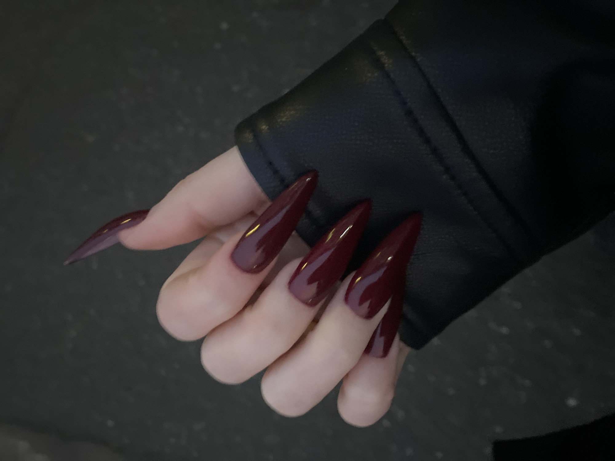 A close-up shot features a hand with elongated, pointed nails painted in a glossy, dark burgundy color. The hand is adorned with a black leather jacket. The background appears to be a dark, indistinct surface. The image's location cannot be determined from the provided visual information.
