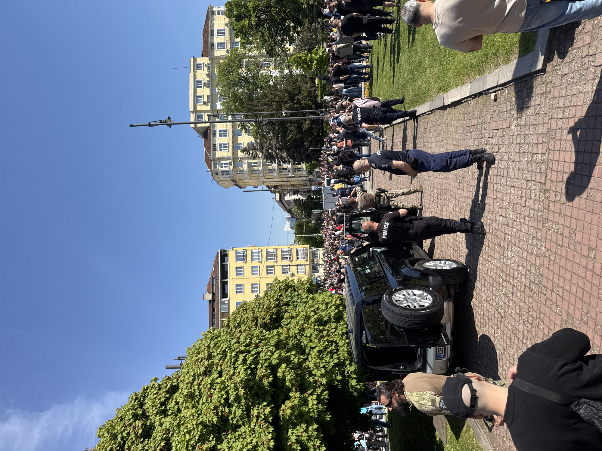 The image depicts an outdoor scene on a brick-paved area in what appears to be the city of Sofia, Bulgaria, on a sunny day. Buildings with yellow facades are visible in the background. A dense green tree stands beside a black SUV with the word "POLICE" visible on the side, and the door is open. Several uniformed police officers are standing near the vehicle, and a crowd of people is gathered further down the pathway near a grassy area and a concrete edge. The scene suggests a public event or gathering that is being monitored by law enforcement.