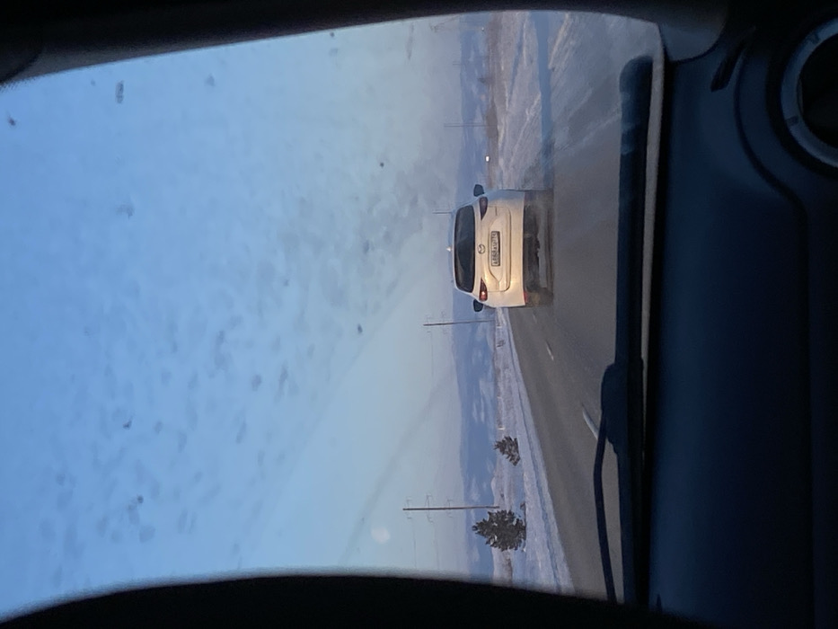 A view from inside a car looking out onto a snowy road. A white car with its license plate visible is directly ahead, traveling in the same direction. Snow covers the ground on either side of the road. The windshield is partially obscured by what appears to be water spots or ice. The car's interior is visible on the sides of the frame, with the dashboard and a partially visible air vent.