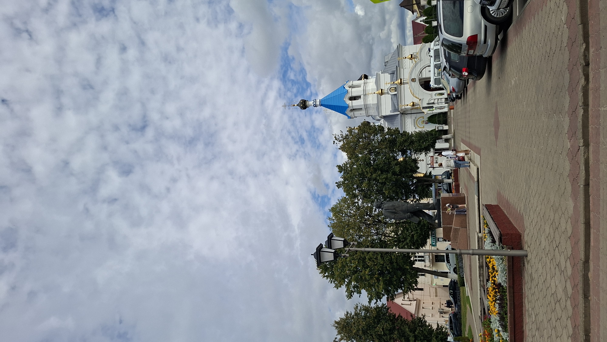 The image shows a view of the Church of the Assumption of the Blessed Virgin Mary in Pastavy, Belarus. A statue of a man is in the foreground, with a lamppost to its left. Several cars are parked in front of the church. People are walking on the sidewalk in front of the church. The sky is cloudy.