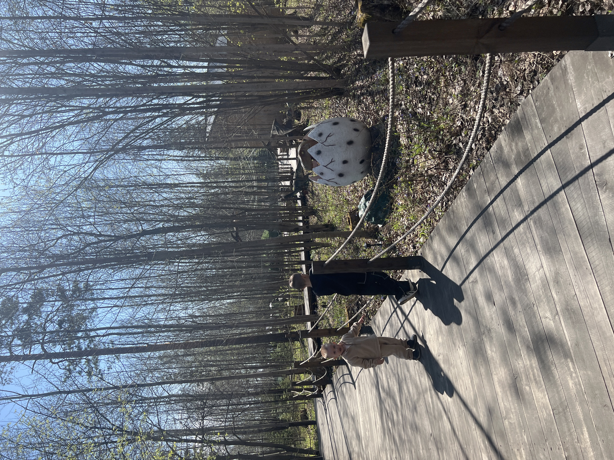 The image depicts an outdoor scene on a wooden walkway, likely in a park or wooded area near Izhevsk, Russia. The perspective is oriented along the walkway. Two individuals are present: an adult and a child. The adult is wearing dark clothing and is standing. The child is wearing light-colored clothing and is also standing. To the side of the walkway, there is a large, cracked, white egg with dark spots, suggesting a dinosaur exhibit. The surrounding environment consists of trees without leaves and ground covered with leaves and vegetation. A rope railing runs along the side of the walkway. The sky is visible through the trees.