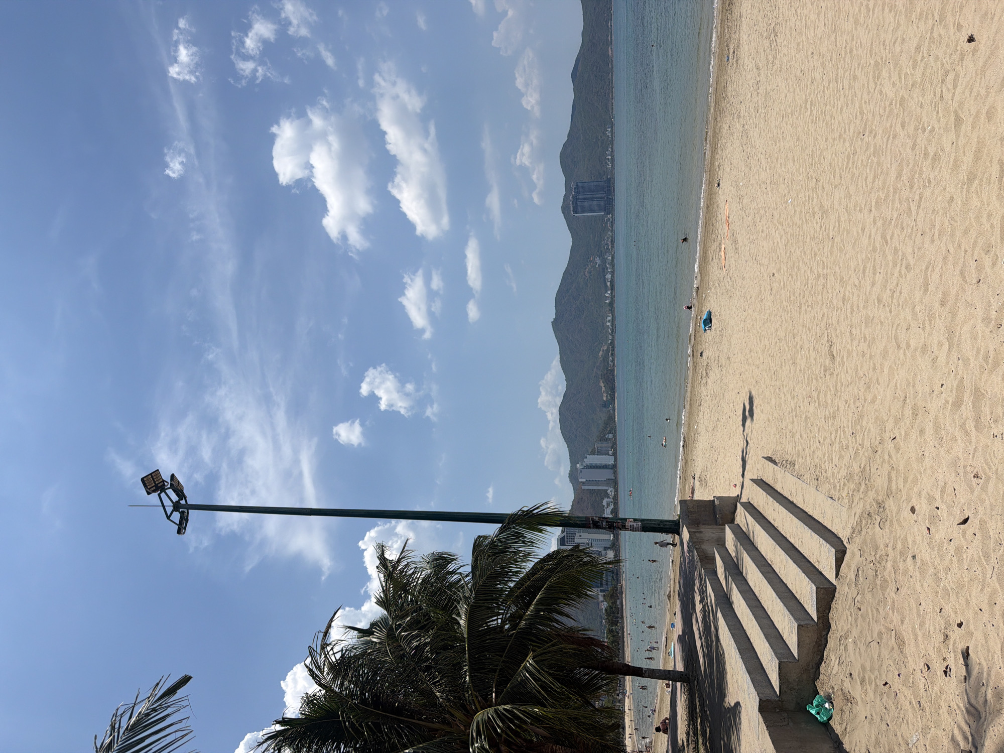 The image depicts a beach in Bac Nha Trang Ward, Vietnam. The scene is taken at a side angle. The beach has tan sand and a gentle slope into the light blue-green water. In the background, there are mountains and buildings. There are some people visible in the water and on the beach.
In the foreground, there is a palm tree and a tall light pole with several light fixtures. A set of concrete steps leads from the beach up to a slightly higher level. The shadows of the steps are cast on the sand.