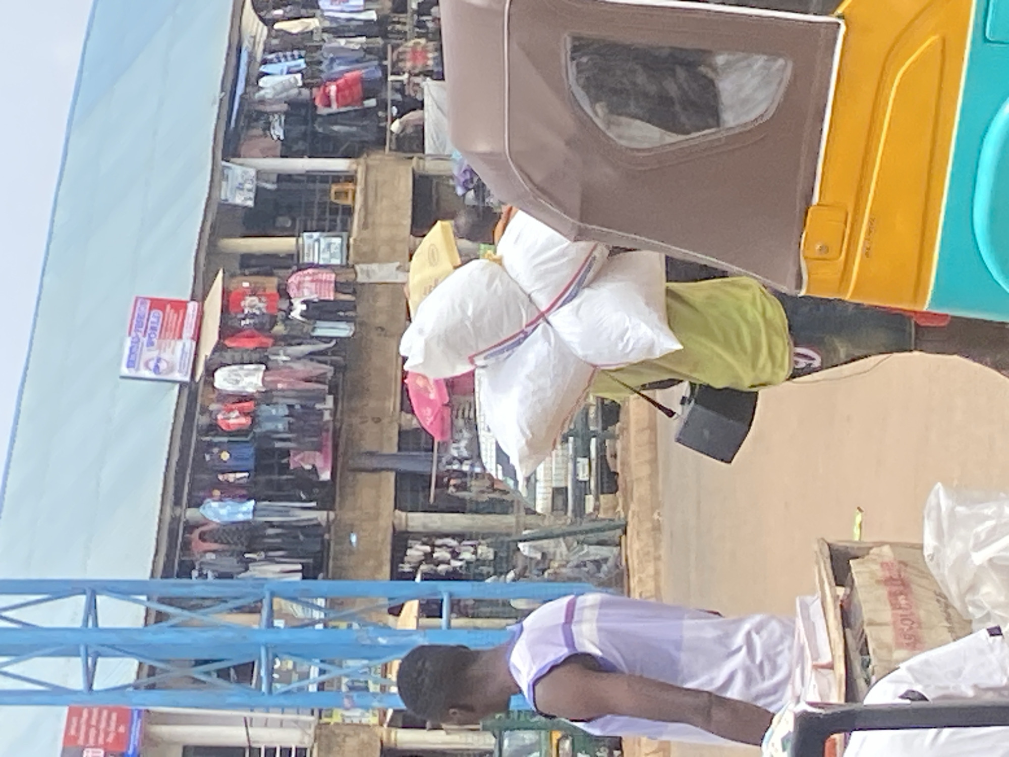The image depicts a bustling street scene in Bukuru, Nigeria. In the foreground, a man wearing a purple and white striped tank top is partially visible on the left. To his right, another man, seen from the back, is carrying a large load of white sacks tied together with red and blue twine. He is also wearing a lime green t-shirt and jeans, and a black rectangular object is hanging from his shoulder. A yellow and turquoise auto-rickshaw is parked in the right foreground.
In the background, a market is in full swing, with numerous clothing stalls lining the street. Clothes of various colors and styles are displayed on racks and hangers. A blue metal structure, possibly part of the market's infrastructure or a nearby building, stands prominently on the left. Signs advertising "SUPER-VALUE WORLD" are visible above the stalls. People can be seen browsing and interacting within the market. The overall environment suggests a lively commercial area in a Nigerian city.