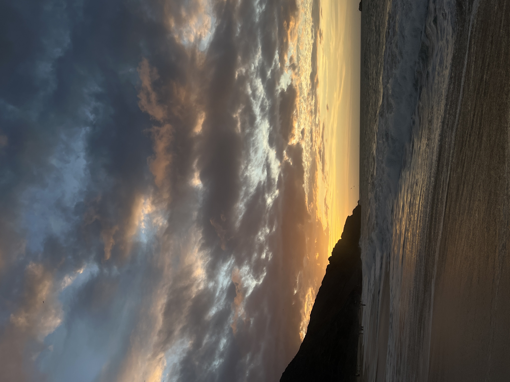 The image depicts a beach scene at dusk, likely in Altagracia, Venezuela. The sky is filled with dark, heavy clouds illuminated by the setting sun, casting an orange and gold glow across the horizon. A dark, silhouetted mountain or large rock formation juts out towards the ocean. The ocean's waves crash onto a sandy beach in the foreground. Some figures can be seen walking along the shoreline. The photograph is shot from a low angle.