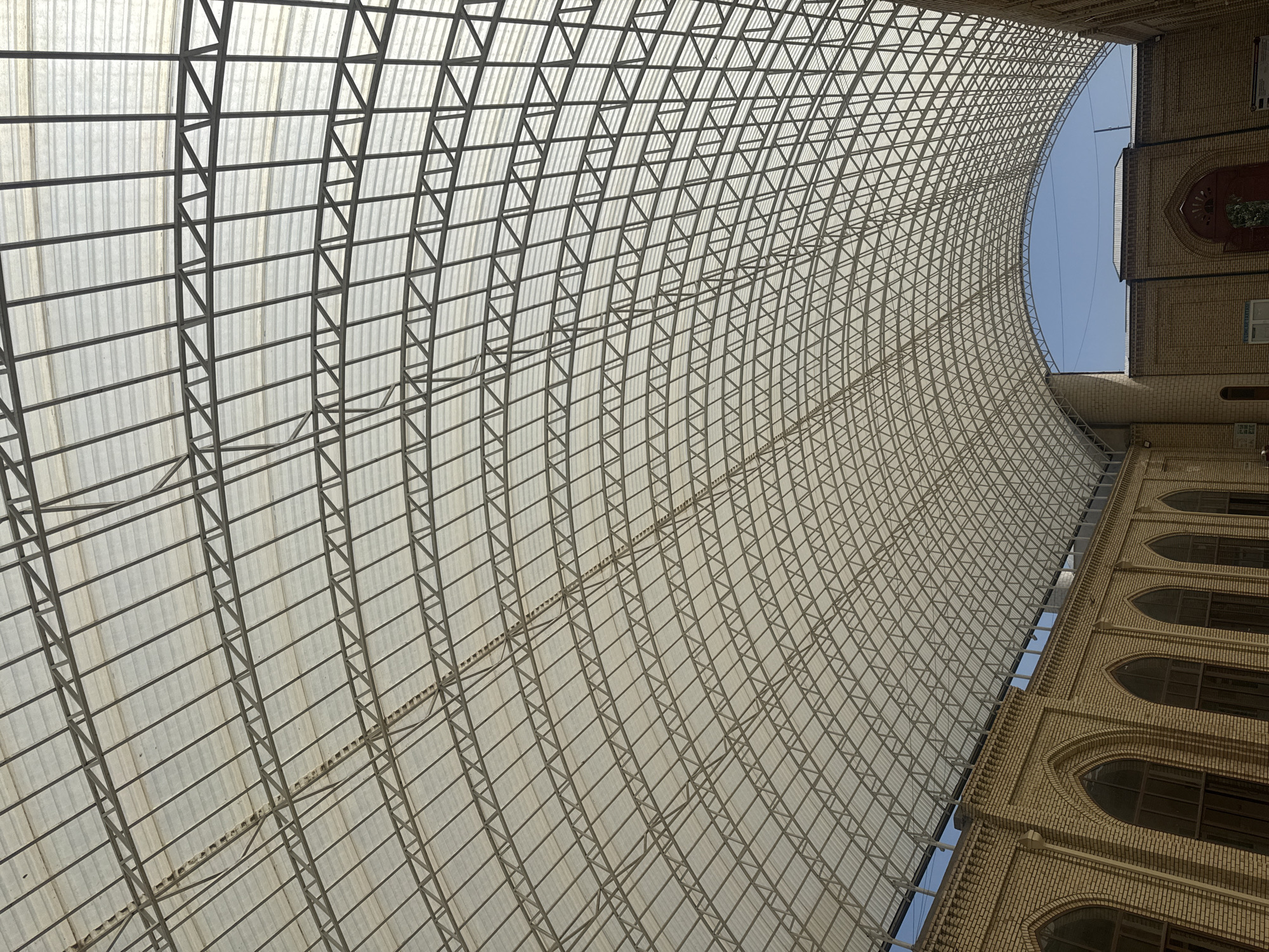 The image shows the interior of a large, arched structure with a translucent roof supported by a complex metal framework. The roof is composed of a grid of metal beams and trusses, overlaid with corrugated translucent panels. Below the roof, the walls of the structure are made of light brown brick, with arched windows and architectural details. The scene is brightly lit by natural light filtering through the roof. The sky is visible through the arched opening of the roof. This structure is likely a covered market or a large public building in Yangikurgan, Uzbekistan, given the architectural style of the brickwork. There are no people visible in the image.