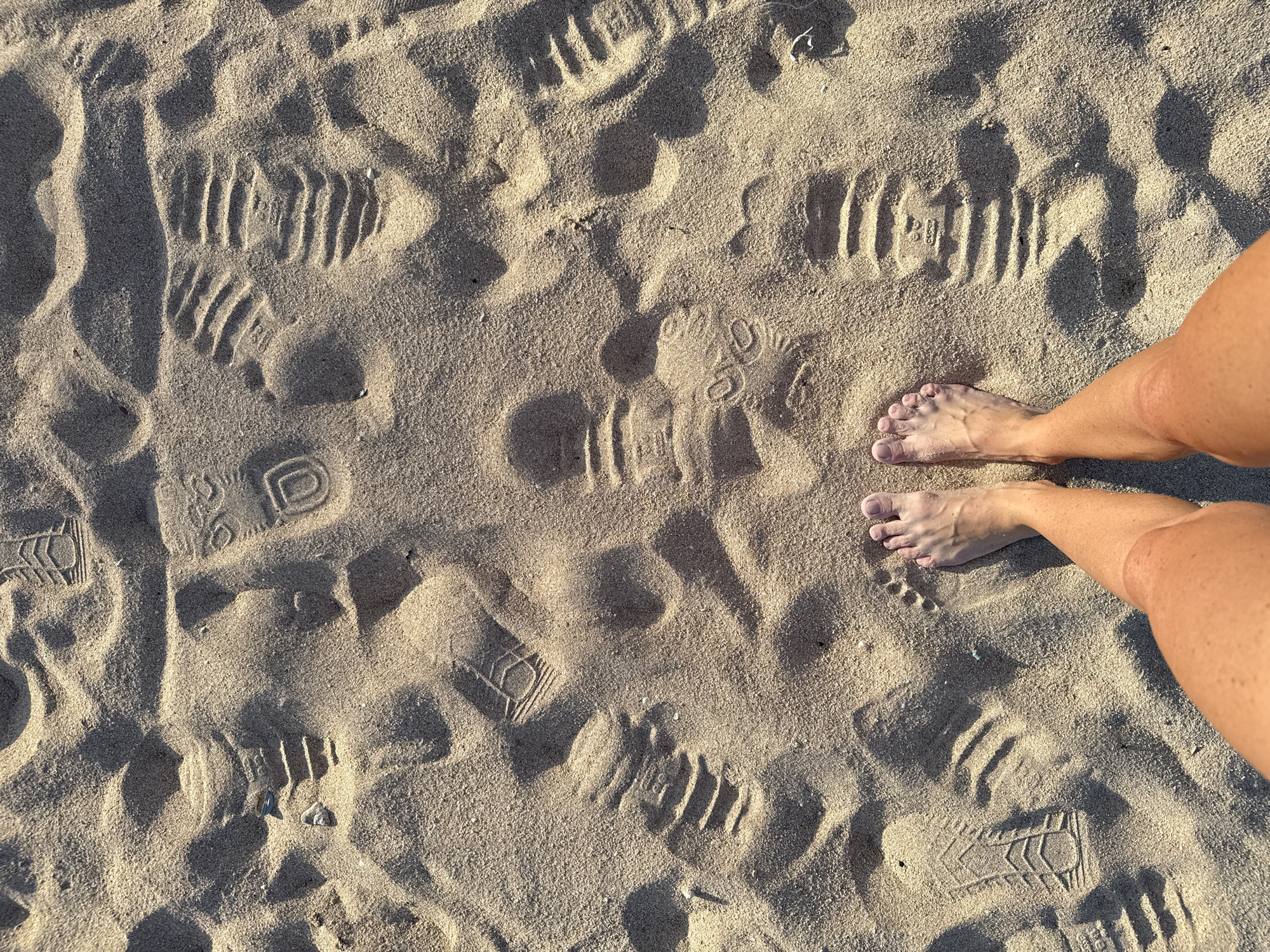 The image shows a sandy beach in Kuta Selatan, Indonesia. The sand is light brown and has numerous footprints, suggesting previous foot traffic. The footprints exhibit different patterns, some with parallel lines and others with more intricate designs that look like tread marks from shoes. In the lower right corner are the bare feet and lower legs of a person, partially obscuring the sand and footprints beneath. The lighting indicates it is daytime, with shadows suggesting the sun is positioned overhead.