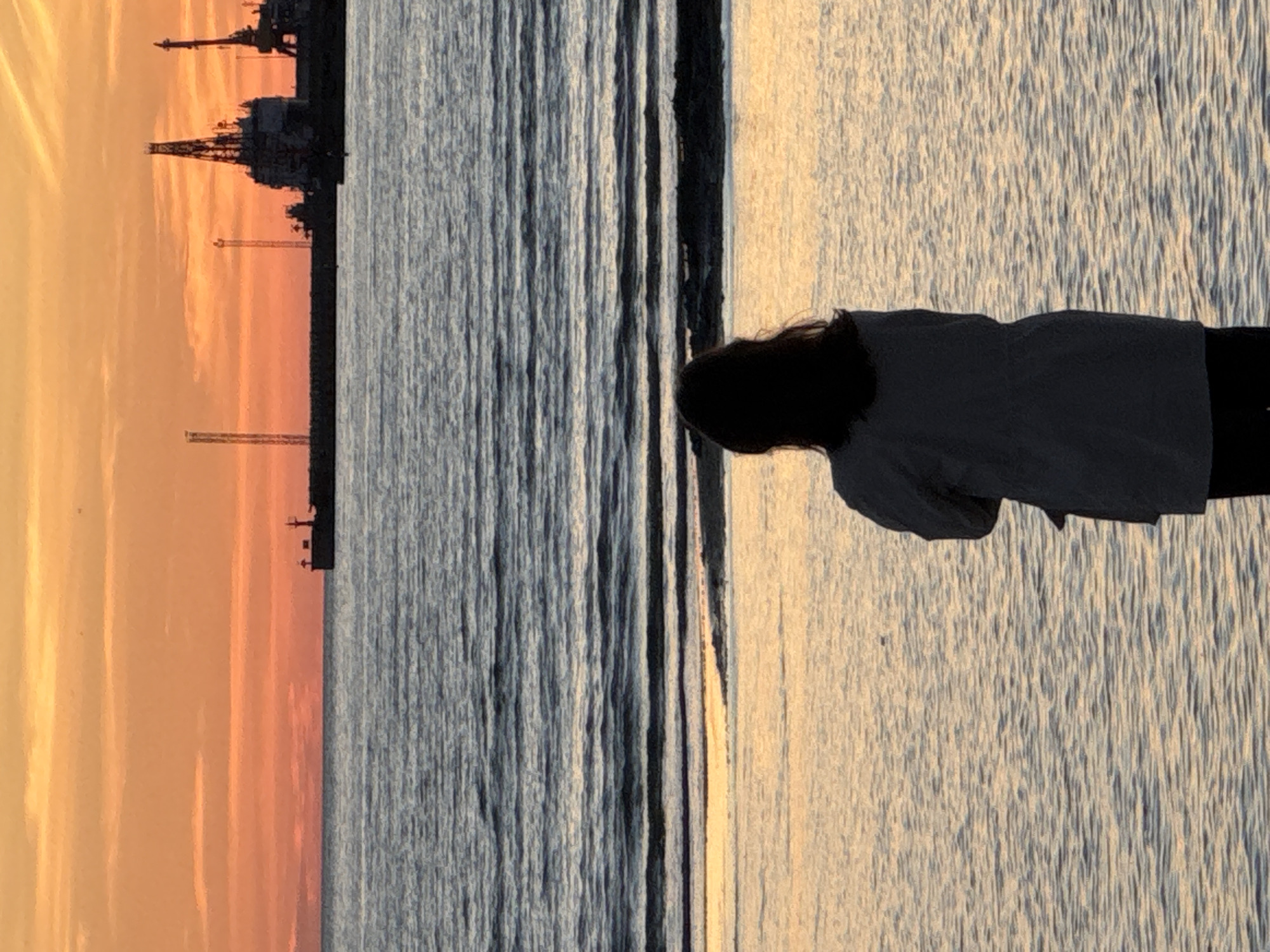 An individual stands on a beach at sunset, facing the water. The sky is a vibrant gradient of orange and yellow, with wispy clouds. The sea is a calm blue with gentle waves. In the distance, the silhouettes of industrial structures and ships are visible against the horizon. The person is wearing a jacket and dark pants, with their back to the camera. The scene is peaceful and serene, with the setting sun casting a warm glow over the water. The industrial silhouette in the background could suggest a port or industrial area, fitting for a coastal city.
