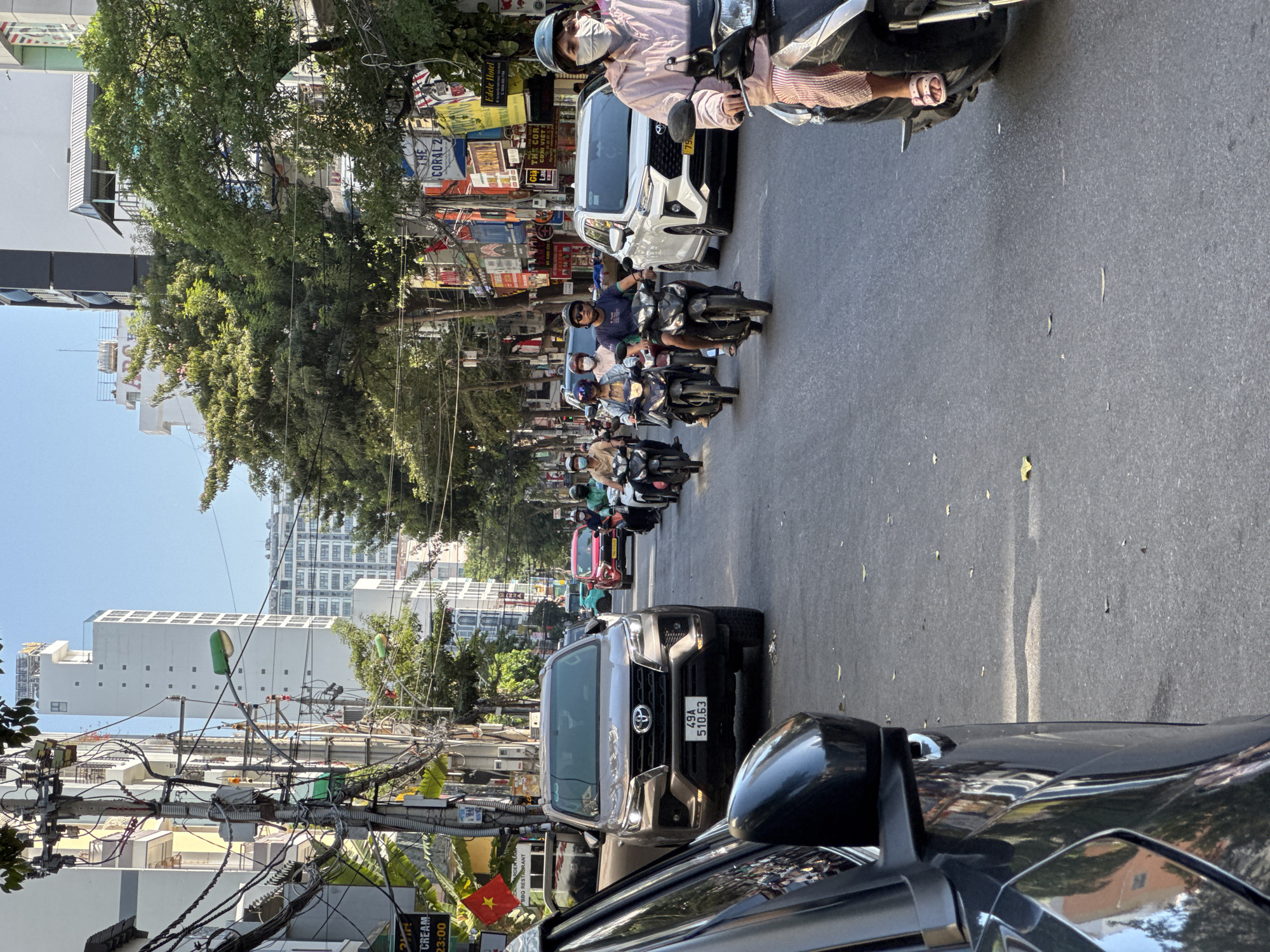 This is a street-level, eye-level, medium shot of a busy street in Nha Trang, Vietnam, on a sunny day. The scene is dominated by a significant amount of electrical wiring overhead, characteristic of urban Vietnamese streets. In the foreground, on the left, the side of a dark-colored SUV is visible. The main focus is on a convoy of motorcycles moving down the street.
Several individuals are riding motorcycles, most wearing helmets and some wearing face masks. A woman in the foreground on a motorcycle is wearing a pink long-sleeved shirt and striped pants, with pink sandals. She has a face mask on and is looking forward. Behind her, other motorcyclists are visible, including a group of three moving together. A white SUV is also present in the middle ground, to the right of the central lane.
The street is asphalt, with some scattered leaves. Buildings line both sides of the street, featuring a mix of modern and older architecture, with balconies and signs. The presence of tall buildings and dense urban infrastructure suggests a commercial or residential area. The overall impression is one of vibrant, daily life in a bustling Vietnamese city.