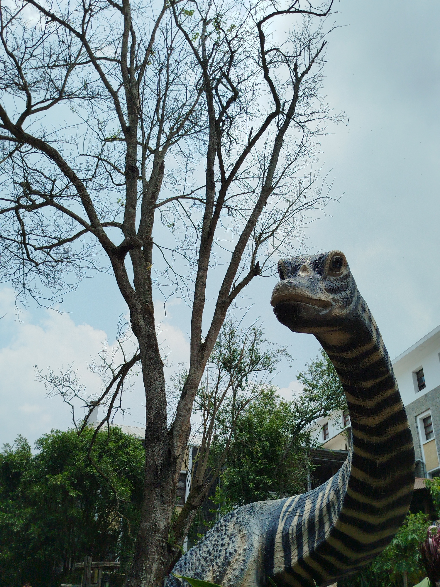 A low-angle shot depicts a dinosaur statue in front of a bare tree, with buildings and greenery in the background. The dinosaur has a long neck and a striped pattern. The location is Lang Biang - Đà Lạt, Vietnam. The sky is partly cloudy.