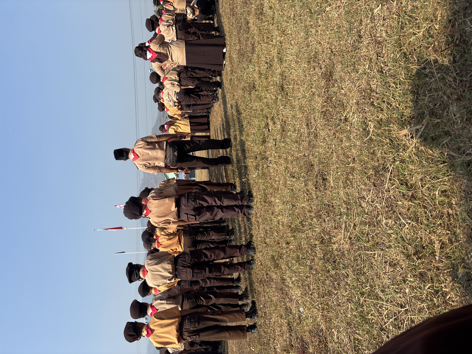 In the city of Tirtomoyo, Indonesia, a group of scouts, predominantly male and wearing brown uniforms with red neckerchiefs, are assembled in a grassy field. They are standing at attention, facing away from the camera. Several flags, including the Indonesian flag, are visible in the background, suggesting an official ceremony or event. The sky is clear and blue, indicating fair weather. The scene is likely a formal gathering, possibly a flag ceremony or a scout jamboree, taking place outdoors in an open area.