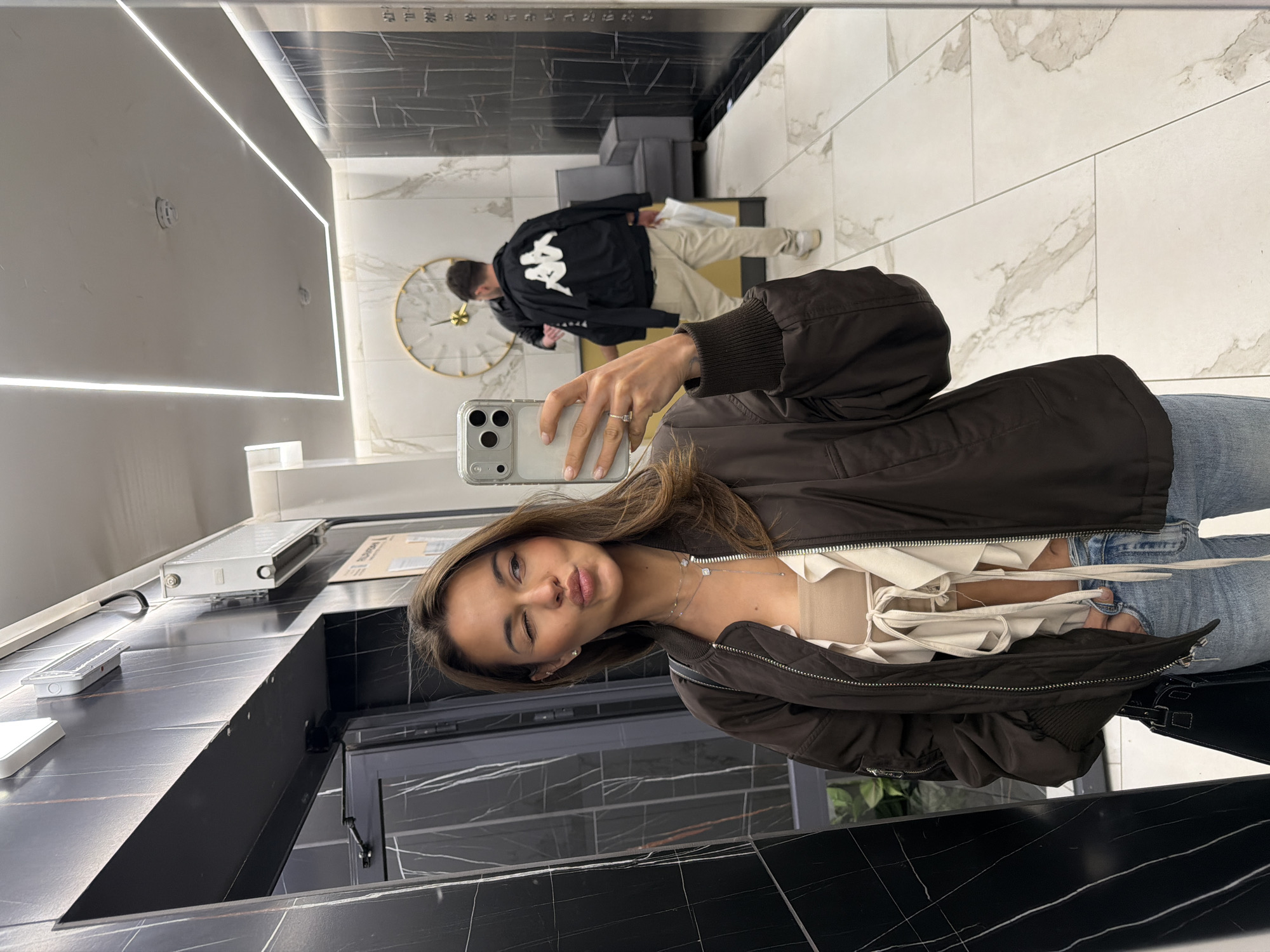 A young woman captures a mirror selfie in what appears to be a modern, stylish interior, possibly a hotel lobby, showroom, or a high-end public space in Krasnoyarsk, Russia. The image is rotated 90 degrees clockwise.
The woman, the primary subject, is positioned in the foreground, holding a light-colored iPhone with a clear case to take the photo. She has long brown hair, wears a dark brown bomber jacket, a light cream-colored top with a ruffled design, and blue jeans. She winks playfully at the camera, with a slight pout on her lips. A delicate silver necklace adorns her neck, and a silver ring is visible on her left ring finger.
Reflected in the background behind her is a man wearing a black jacket with a prominent white "KAPPA" logo on the back, and light beige pants. He appears to be standing or walking, facing away from the mirror.
The setting features sleek, contemporary design elements. The walls and floor are covered in large, polished tiles, with a distinct black marble pattern on the wall/counter behind the woman, and a lighter white/grey marble pattern on the floor/wall behind the man. The ceiling is plain white with modern, linear LED light strips running along the edges. A gold-framed wall clock with gold hands is visible on the wall behind the man. On the dark marble counter to the woman's left, there's a white radiator, a cardboard box, and a modern pop-up electrical outlet. Dark, minimalist furniture (possibly chairs or a sofa) can be seen further in the background. The space is brightly lit, suggesting it's either daytime or a well-lit interior at night. The overall impression is one of sophistication and contemporary style.