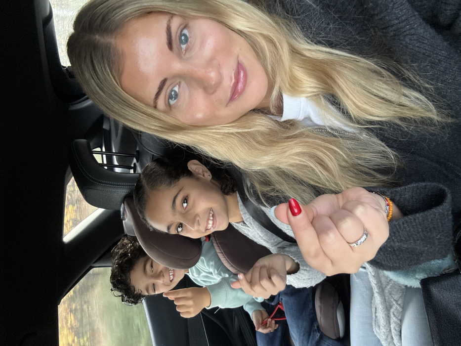 The image depicts three individuals inside a vehicle located in Pourrières, France, during daylight hours. In the foreground, an adult female with long, wavy blonde hair, light skin, and blue eyes is visible, facing slightly towards the camera. She wears a white high-neck top under a dark grey zippered cardigan. Her right hand is raised, forming a "finger heart" gesture, with the thumbnail painted red. A ring is present on her right ring finger, and beaded bracelets are visible on her right wrist. She displays a faint smile.
In the rear passenger area, two children are seated in booster seats. On the right, a girl with dark, pulled-back curly hair, light brown skin, and dark eyes, wearing a grey long-sleeved top, smiles directly at the camera. To her left, a boy with dark, curly hair, light brown skin, and dark eyes, wearing a light blue long-sleeved top and jeans, also smiles directly at the camera while performing a "finger heart" gesture with his right hand. A red object is partially visible near the boy's lap. The car's interior is dark, and blurred outdoor scenery, characterized by yellowish-brown foliage, is visible through the rear window.