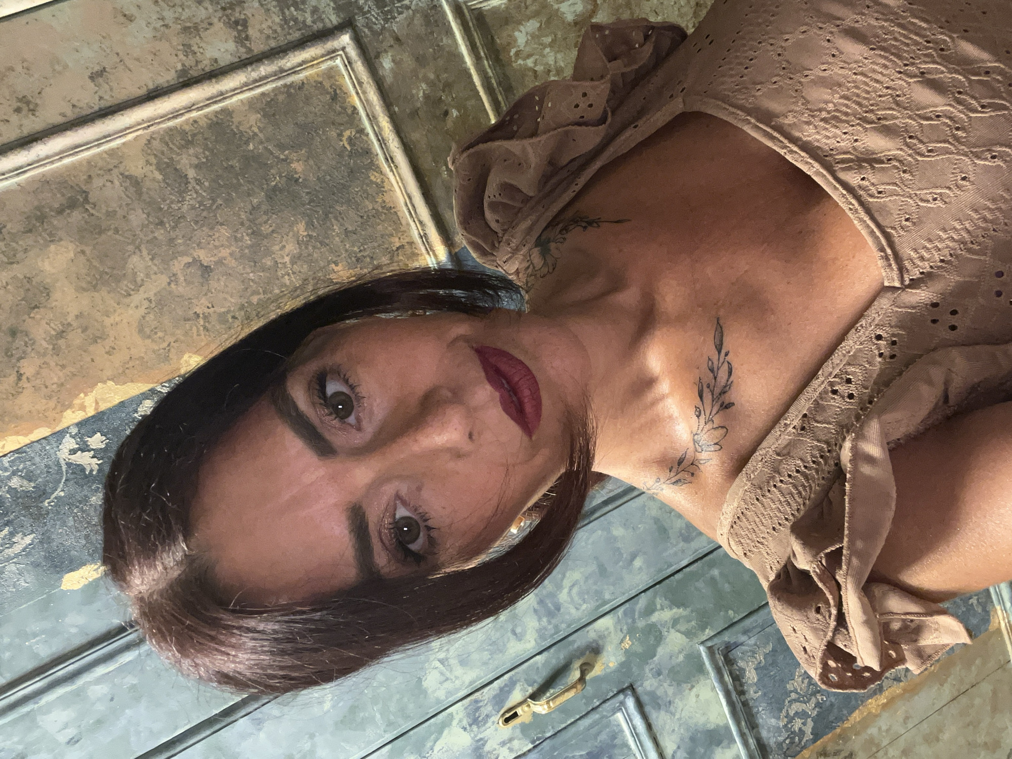 An extreme close-up selfie shows a woman with medium-length dark hair, fair skin, and dark eyes. She is wearing a light brown, textured top with ruffled sleeves and decorative cutouts. A tattoo of a floral branch is visible on her upper chest and another on her collarbone. The background appears to be an aged, textured wall with hints of green and gold. The overall impression is a selfie taken indoors, possibly in an artistic or antique setting. The provided location information is not consistent with the visual content of the image.