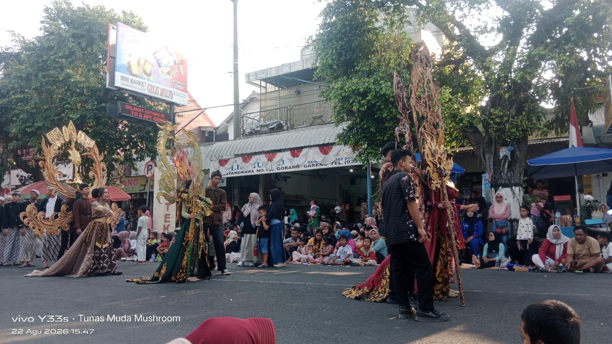 This image depicts a street scene in Genengan, Indonesia, during what appears to be a cultural event or parade. The foreground features several individuals in elaborate traditional costumes, adorned with golden, intricate headdresses and flowing garments. Two particularly prominent figures are adorned with large, golden, winged structures, one resembling a dragon and the other a more abstract floral or sun-like design.
Behind these main subjects, a crowd of spectators is gathered on the side of the street, many seated on the ground. They are dressed in a variety of casual clothing, with some individuals wearing headscarves. The background shows a street lined with buildings, some with awnings and shops. A large tree with dense foliage is visible on the right side of the frame, and a blue umbrella provides shade for some of the spectators. Signs are visible in the background, one of which appears to be a directional or informational sign with Indonesian text. The overall lighting suggests it is daytime, possibly late afternoon, given the angle of the sunlight. The image also contains a watermark at the bottom left corner indicating "vivo Y33s · Tunas Muda Mushroom 22 Agu 2026 15.47," which is a camera model and date/time stamp.