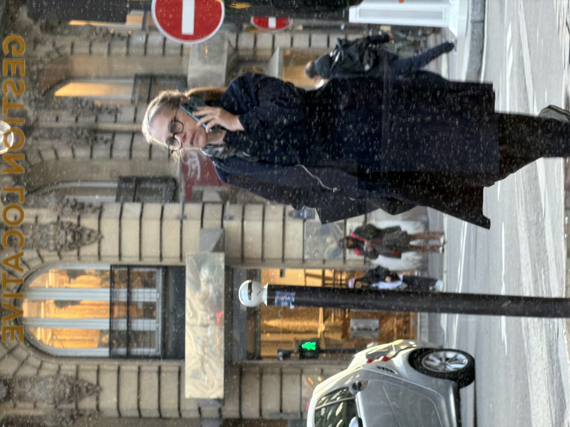 This image depicts a bustling urban street scene in Paris, France, characterized by its classic Haussmannian architecture visible in the background building with ornate stone facades, arched windows, and decorative railings. The location is further indicated by the reversed reflection of "GESTION LOCATIVE" (Rental Management) on an upper window.
In the foreground and mid-ground, a woman in her 40s or 50s, with light hair pulled back and wearing glasses, a dark patterned or textured long coat, and a scarf, walks from left to right. She is actively engaged in a phone call, holding a dark-colored smartphone to her right ear.
On the left side of the street, a silver Smart Fortwo car is partially visible, either parked or in motion. A prominent black street pole with a white spherical top and attached stickers stands in the immediate left foreground. Behind it, a green pedestrian "walk" signal is illuminated.
The background features the detailed stone building with a warm, illuminated commercial entrance at ground level. Several other pedestrians are present: one person wearing a pink hat near the building entrance, another individual in a dark jacket with a red vest appearing to look at a device, and a third person with a black backpack walking away on the right. A clear red circular "no entry" sign with a white horizontal bar is visible on the right side of the frame. White pedestrian crossing markings are visible on the asphalt in the lower portion of the image.
The entire scene is viewed through a reflective surface, creating numerous small, blurry, light-colored specks across the image, consistent with rain, snow, or condensation on the lens or glass barrier.