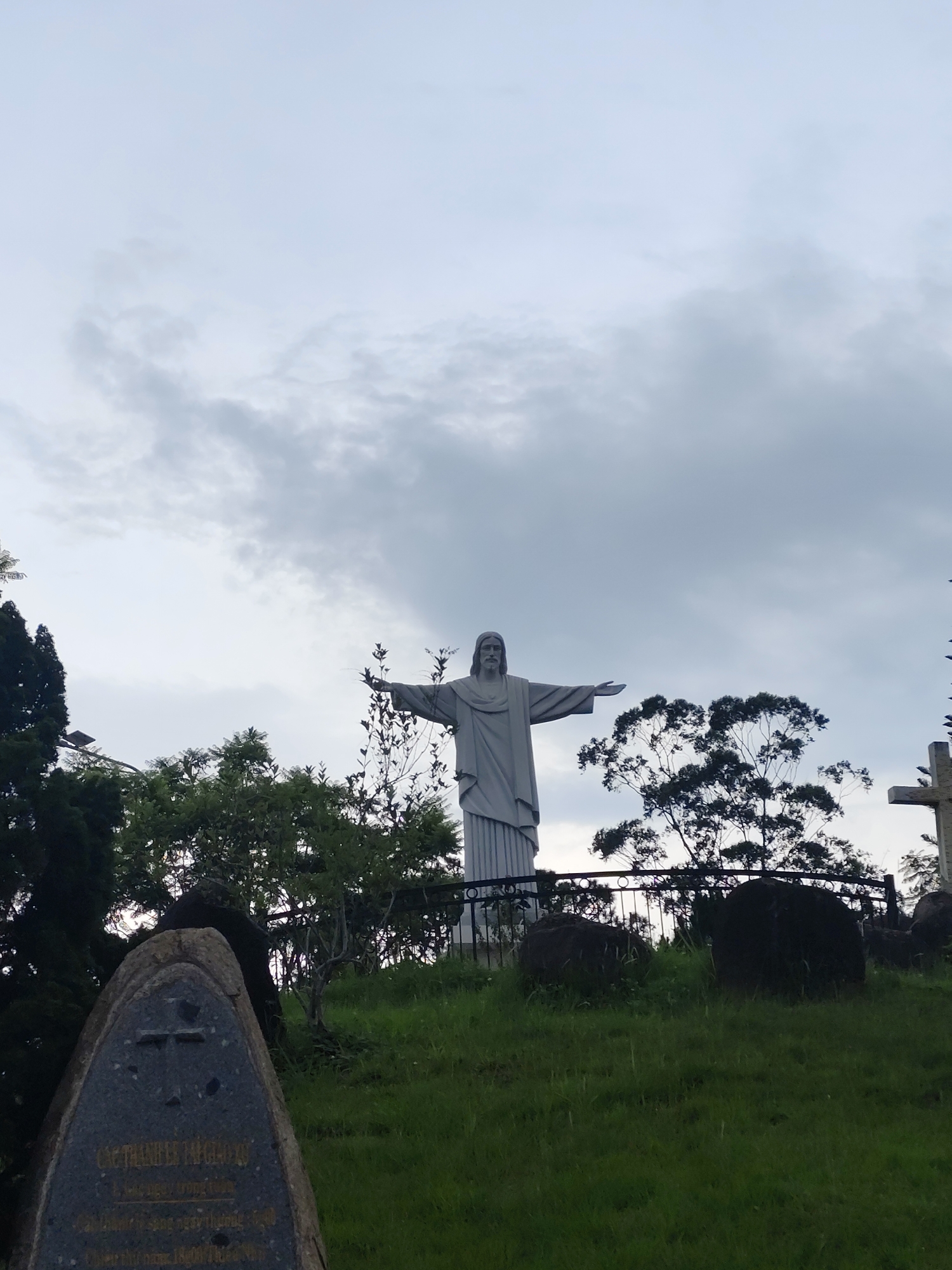 A statue of Jesus Christ stands on a hill in Lạc Dương, Vietnam, with his arms outstretched. The statue is made of light gray stone. The hill is covered in green grass and a few trees. A stone monument stands to the left of the statue and in front of it. Behind the statue is a gray, cloudy sky.