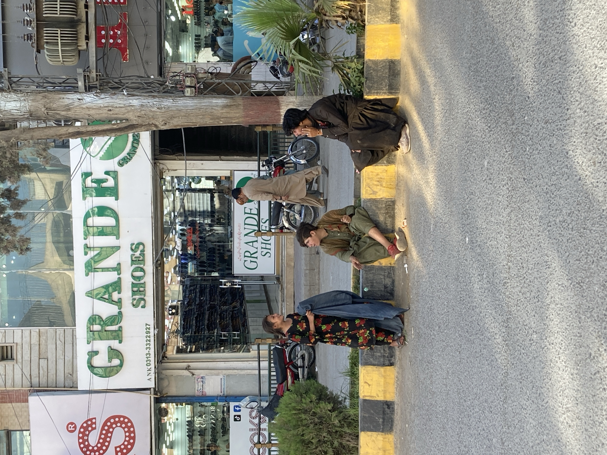 This street-level image captures a bustling scene in what appears to be a commercial area of Quetta City Tehsil, Pakistan, during the late afternoon or early evening. The warm light casts long shadows across the asphalt road and sidewalk.
In the foreground, several individuals are resting or engaging in activities on the sidewalk next to a curb painted with black and yellow stripes. To the right, a young man in a black shalwar kameez is crouched, seemingly talking on a mobile phone with a slight smile. Next to him, another person in a green outfit is seated on the curb, looking down. Closer to the left, a woman wearing a black floral-patterned dress stands, holding a denim jacket, and appears to be observing the scene to her right. Further back, another man in a light brown shalwar kameez walks past a storefront. Several motorcycles are parked along the busy sidewalk.
The background features the storefronts of various businesses. Most prominent is a shoe store named "GRANDE SHOES," identified by large green lettering on a white sign. Below it, a contact number "ANK 0313-3322927" is visible. Another sign displays text in a local script, "زانانہ اور جگا نہ ایمے رنڈورائی دستیاب ہیں," likely advertising the availability of shoes for men and women. Utility poles with a complex network of electrical wires crisscross above the street, and a tree trunk runs vertically through the center-left of the frame. The scene portrays a typical urban environment with people going about their daily routines.