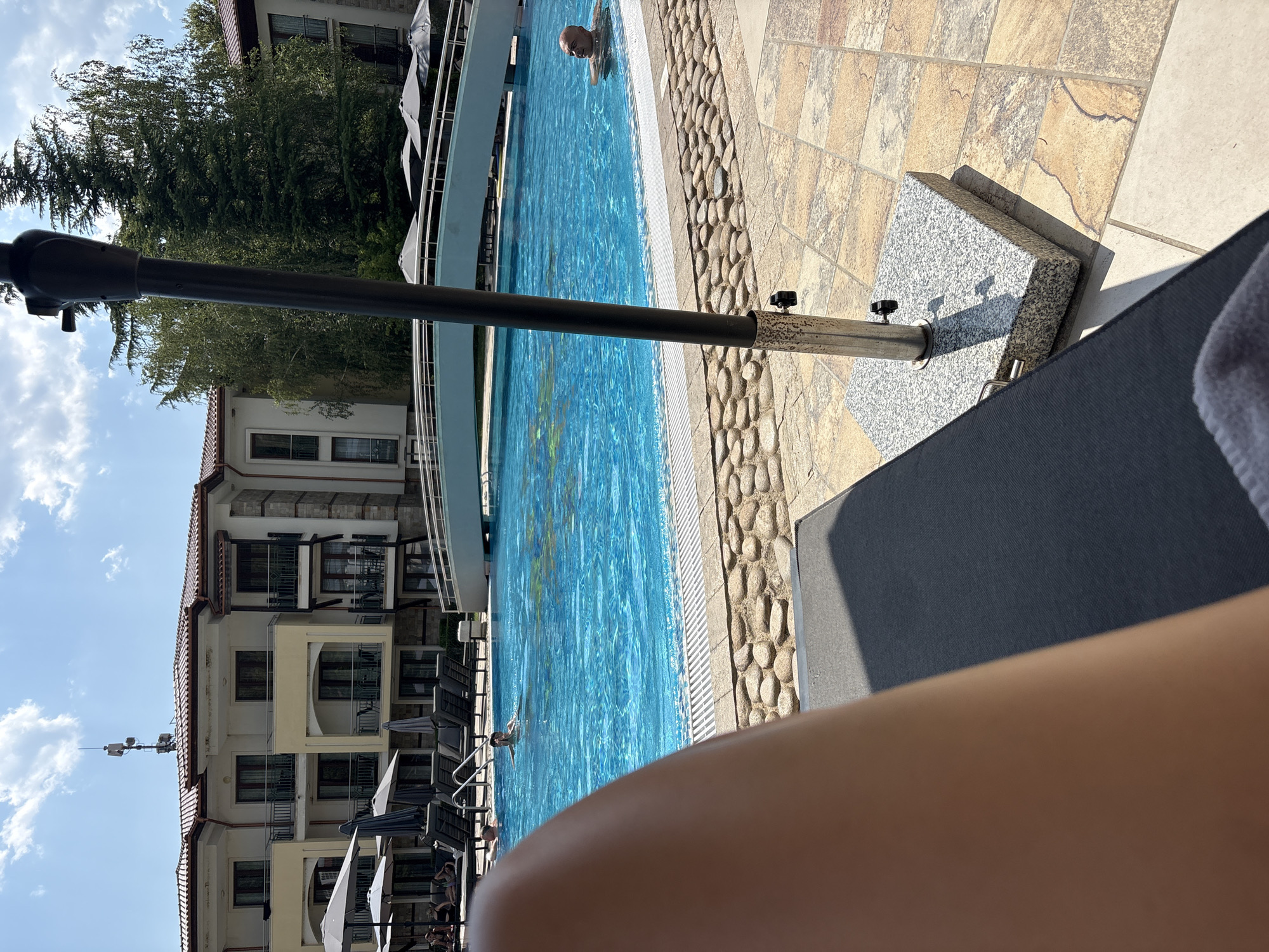 A first-person perspective shot captures a sun-drenched scene at a resort pool. The foreground is dominated by the out-of-focus limb of a person relaxing on a dark gray lounge chair, partially obscuring a granite umbrella stand and a tiled patio. The midground showcases a vibrant blue swimming pool, with several people visible enjoying the water. To the left, a section of the pool is lined with rows of empty lounge chairs and gray umbrellas. In the background, a multi-story building with a terracotta roof and cream-colored facade stands, featuring balconies and stone accents. A white arched bridge spans a portion of the pool. Lush green trees, including a tall pine, rise behind the building. The sky is bright blue with scattered white clouds.