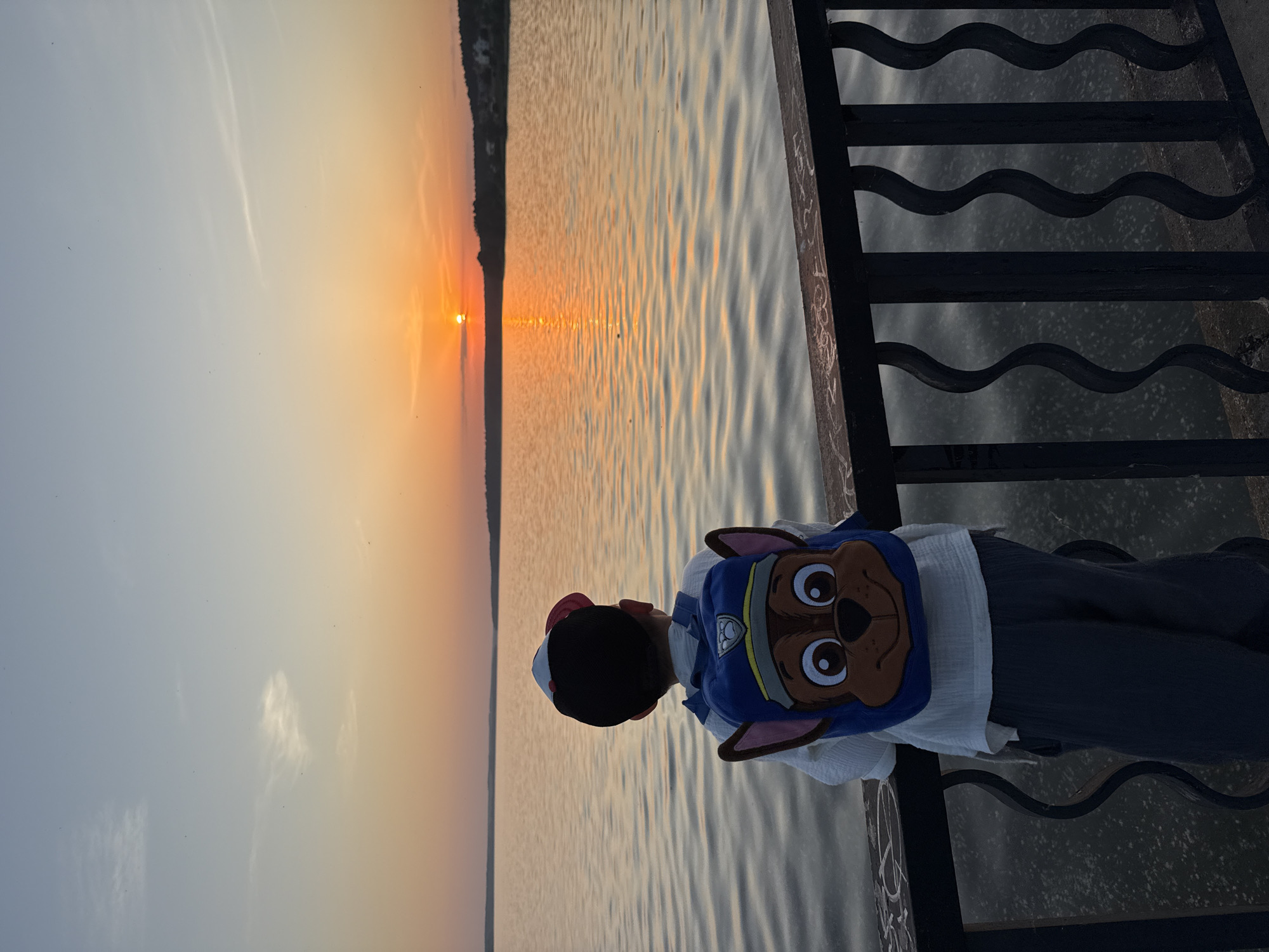 A young child wearing a Paw Patrol-themed hat and backpack stands on a pier in Votkinsk, Russia, gazing out at a lake during sunset. The sun is setting on the horizon, creating a warm, orange glow on the water and the sky. The pier has a black metal railing with a wavy design, and the water is rippled.