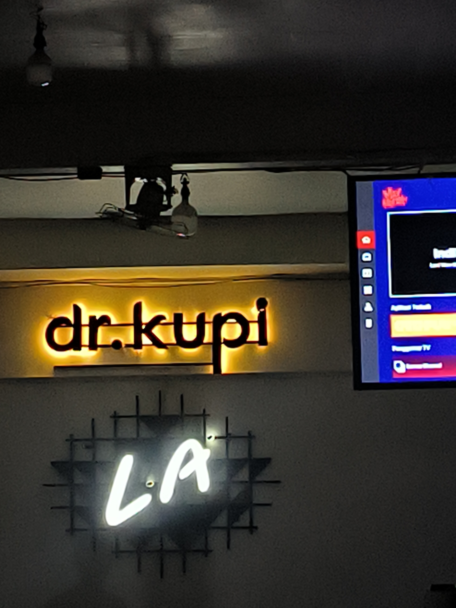 The image shows the interior of a business, likely a cafe, in Lhokseumawe, Indonesia. Above an off-white wall, is a ceiling with two light fixtures. Mounted to the wall is a lighted sign that reads "dr.kupi" in sans-serif letters, above a neon sign which spells out the letters "LA". To the right of the "dr.kupi" sign is a television showing a menu and interface. The general lighting is dim, focusing the viewers attention on the signs and television.