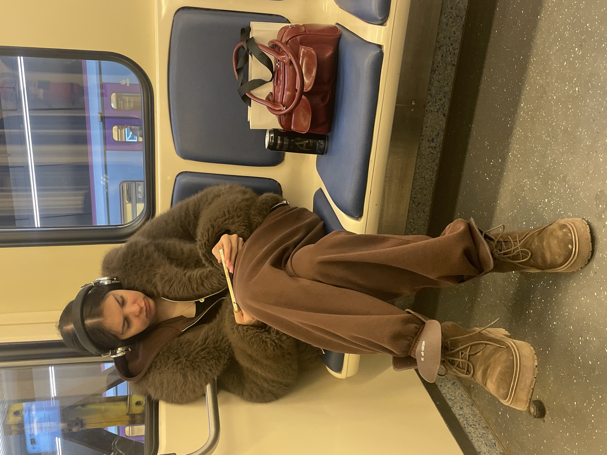 A photograph shows a woman sitting on a train, possibly in Moscow, Russia. The woman is wearing a brown furry coat, brown sweatpants, headphones, and tan-colored boots. She appears to be holding a thin object, possibly a phone or pen. Next to her on the seat is a maroon handbag and a black cylindrical object, likely a beverage container. Reflections of other train cars are visible in the window. The interior of the train is visible with blue seats, yellow panels, and gray flooring with speckled texture.