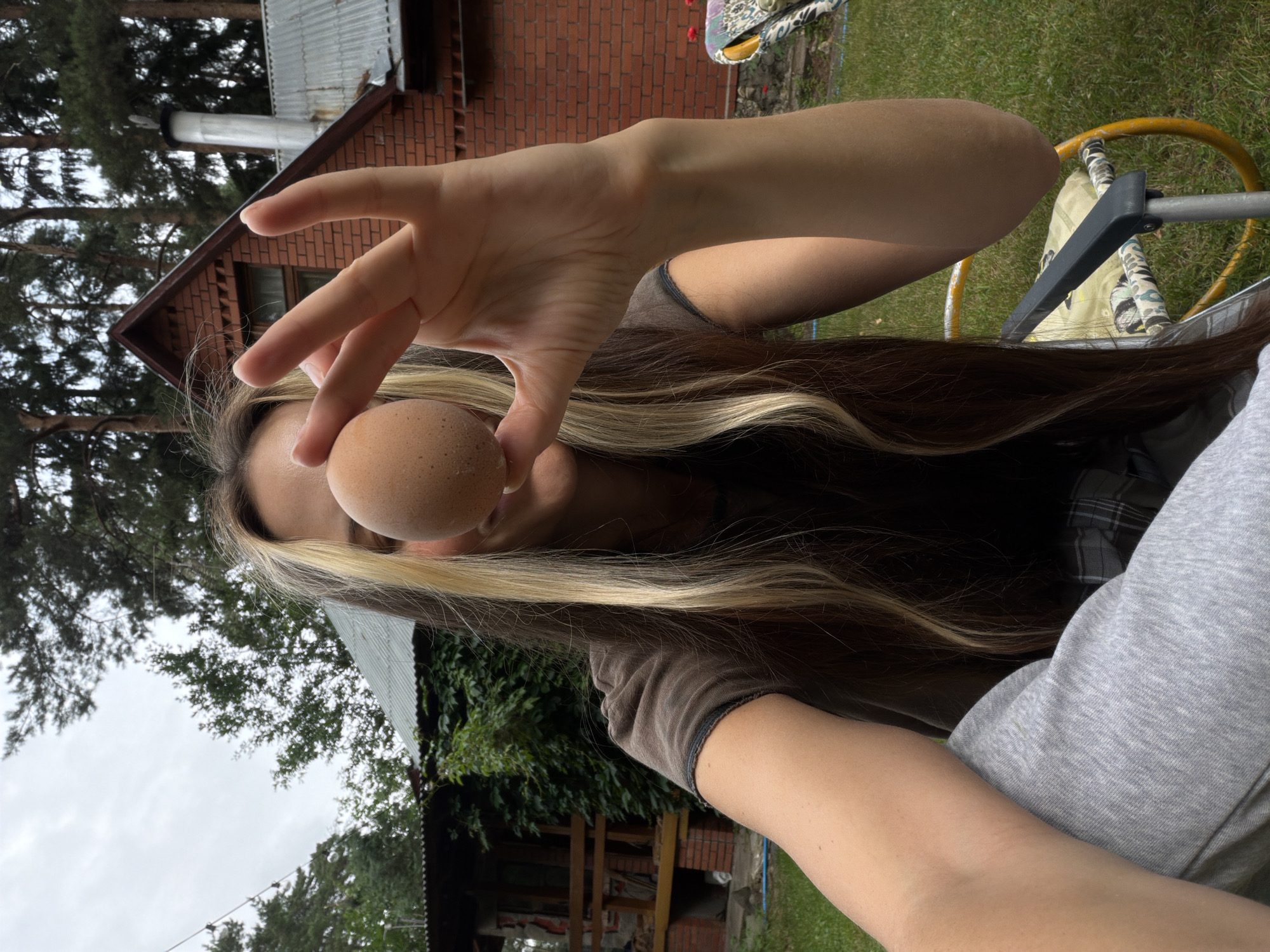 A young woman with long brown hair, featuring blonde streaks, is holding a single brown egg in front of her face, obscuring her eyes and nose. She is wearing a dark brown t-shirt and grey sweatpants. Her right arm is extended, with her fingers forming a peace sign, and the egg is held between her thumb and index finger. She is seated outdoors in what appears to be a backyard. In the background, a red brick house with a corrugated metal roof is visible, surrounded by trees and lush green grass. The setting suggests a rural or suburban environment, consistent with the town of Udelnaya, Russia. The overall atmosphere is casual and relaxed.