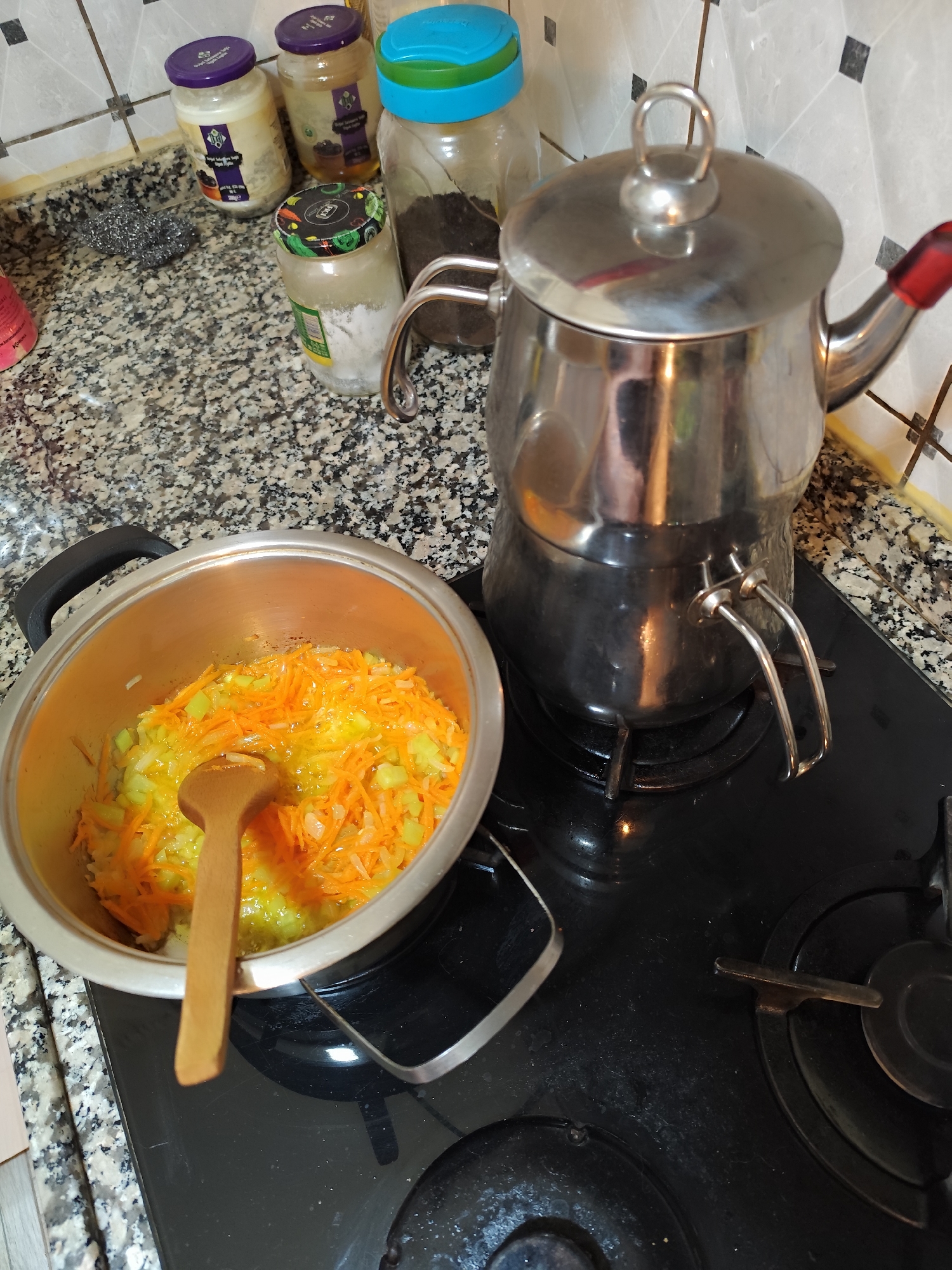A kitchen scene in Sarıyer, Turkey, shows a stainless steel pot filled with a mixture of sauteed orange and yellow vegetables on a black gas stove, a wooden spoon resting in the pot. A two-tiered stainless steel Turkish teapot sits to the right of the pot. Glass jars with various contents are lined up on the speckled countertop behind the pot and teapot. A metal scouring pad is visible on the countertop.